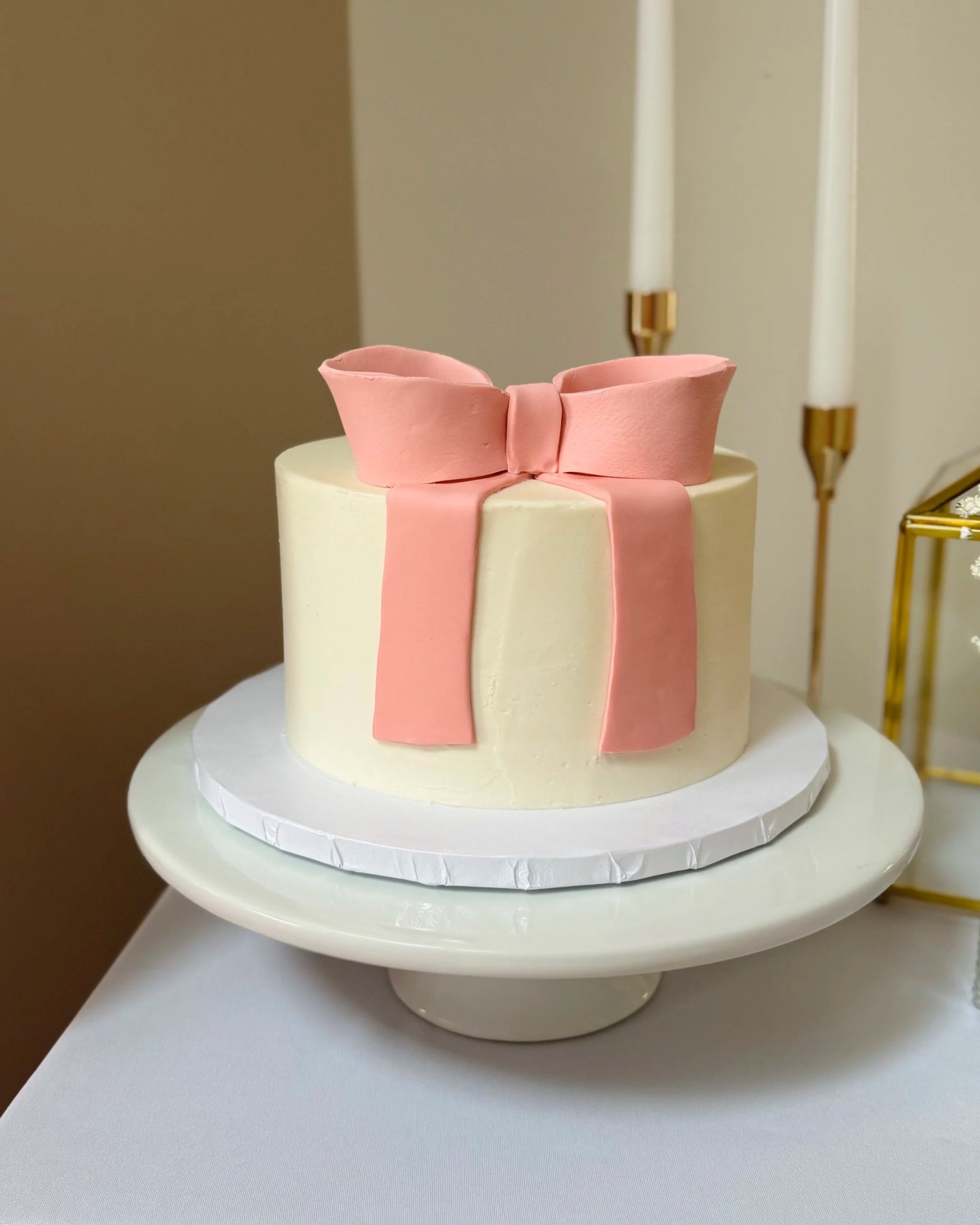 A round white cake decorated with a pink fondant bow on top, placed on a white cake stand.