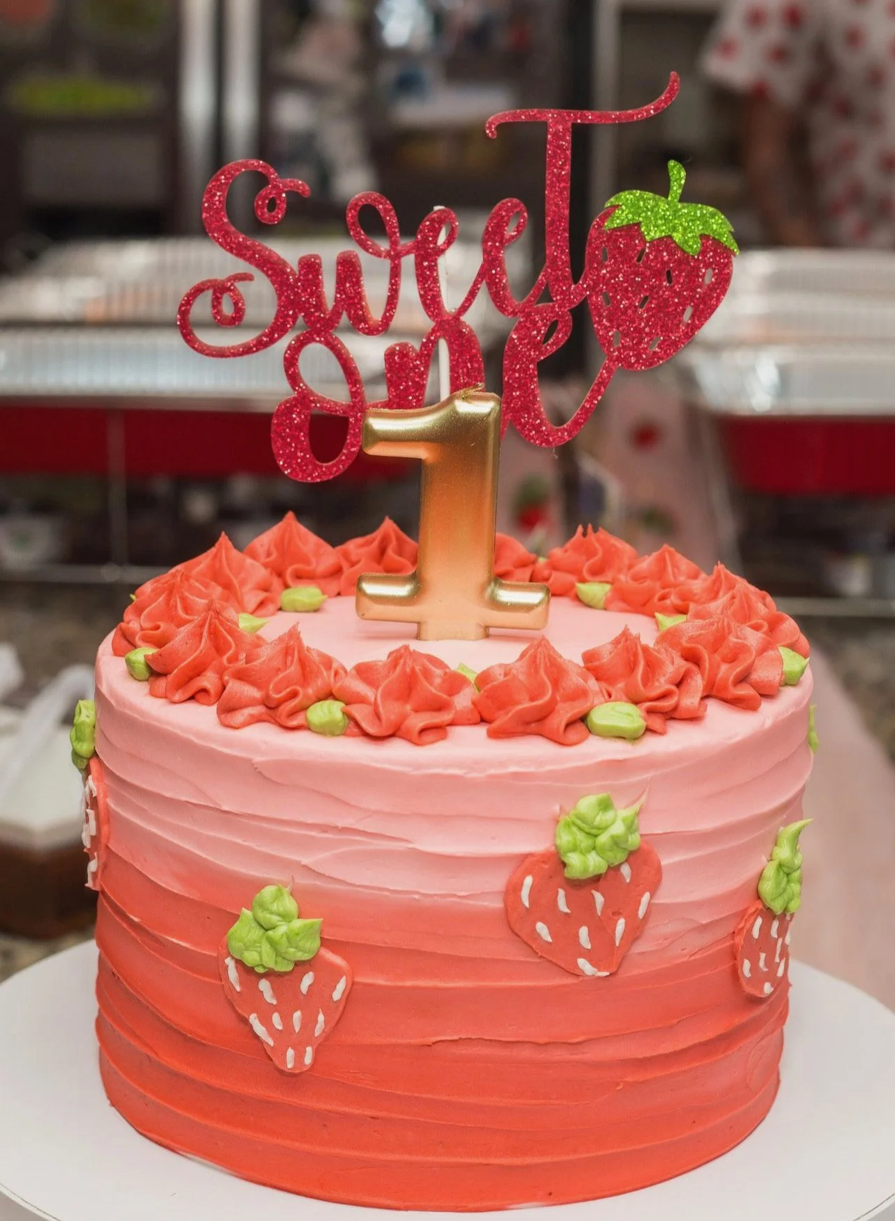 Pink and red strawberry-themed birthday cake with a gold number one candle on top, topped with a red glittery cake topper that says 'Sweet & Tarte' with a strawberry illustration, decorated with small strawberry and green leaf themed icing, set against a blurred background.