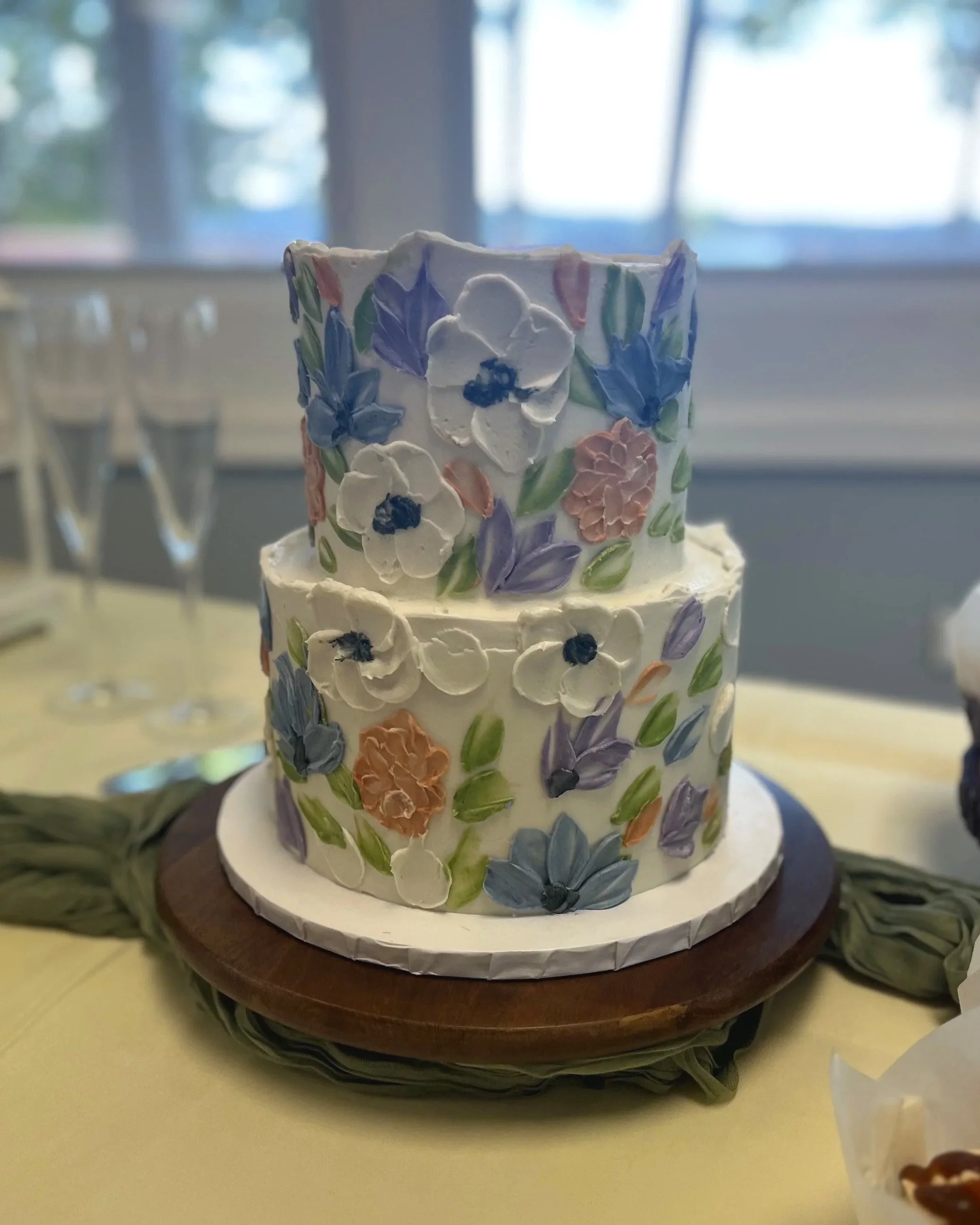 Two-tiered wedding cake decorated with pastel-colored floral icing, sitting on a wooden cake stand on a yellow tablecloth.