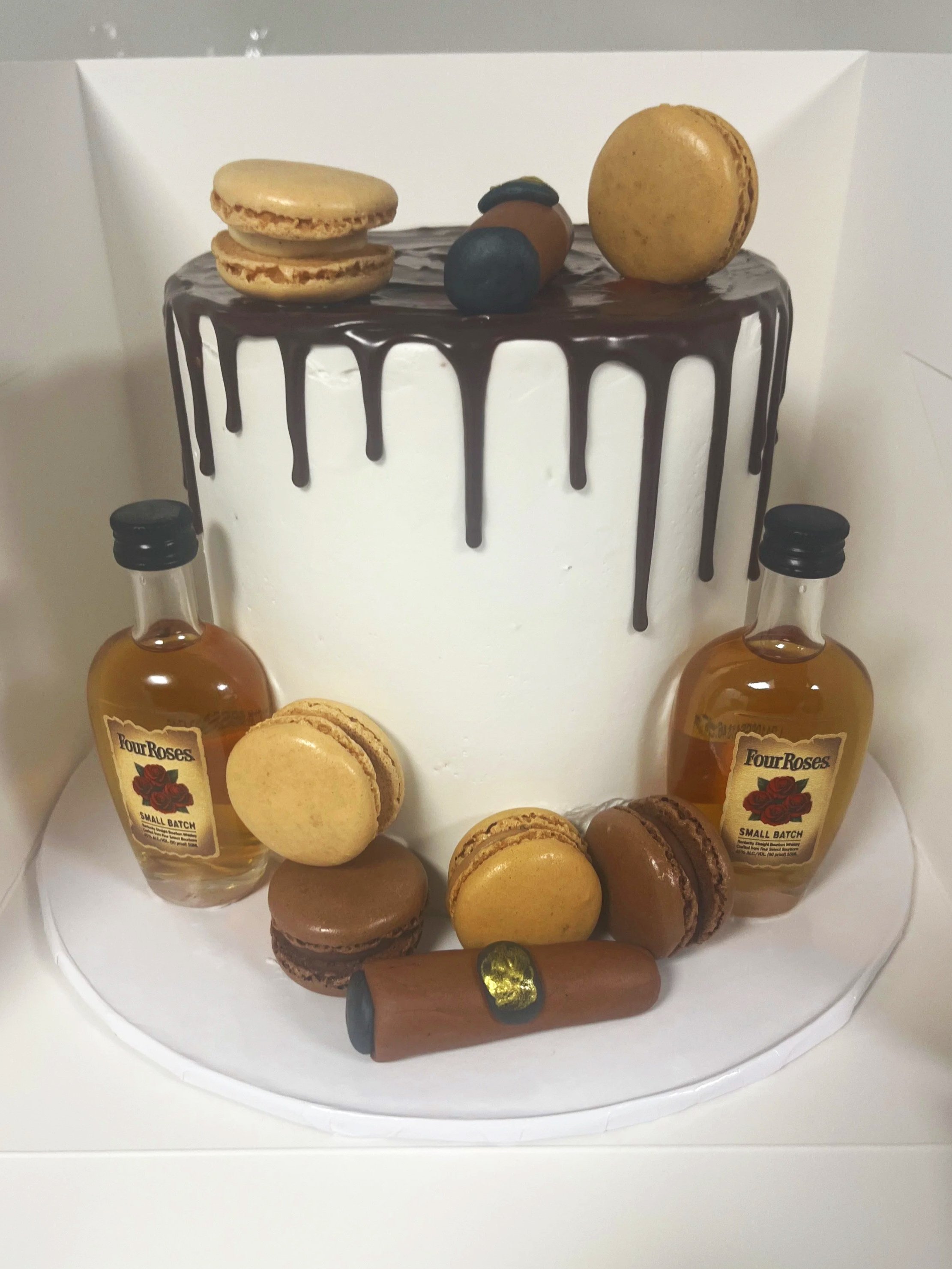 Decorative cake with a white base, topped with chocolate drizzle, macarons, candies, a cigar-shaped pastry, and small bottles of liquor around the base.