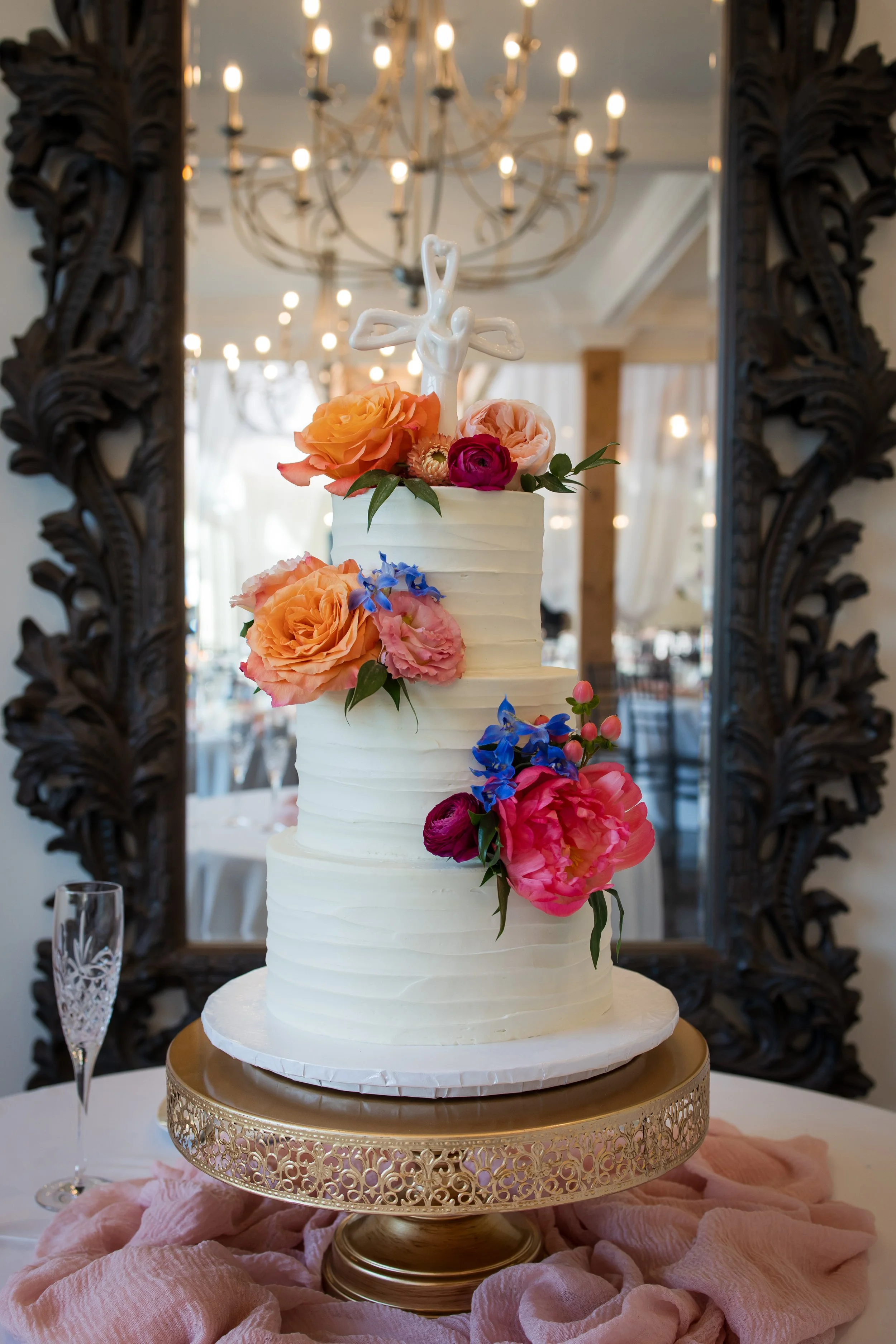 Three-tier white wedding cake decorated with colorful flowers and a white cross topper, placed on a gold cake stand with pink fabric underneath. A champagne glass is beside the cake, with a large mirror and chandelier in the background.