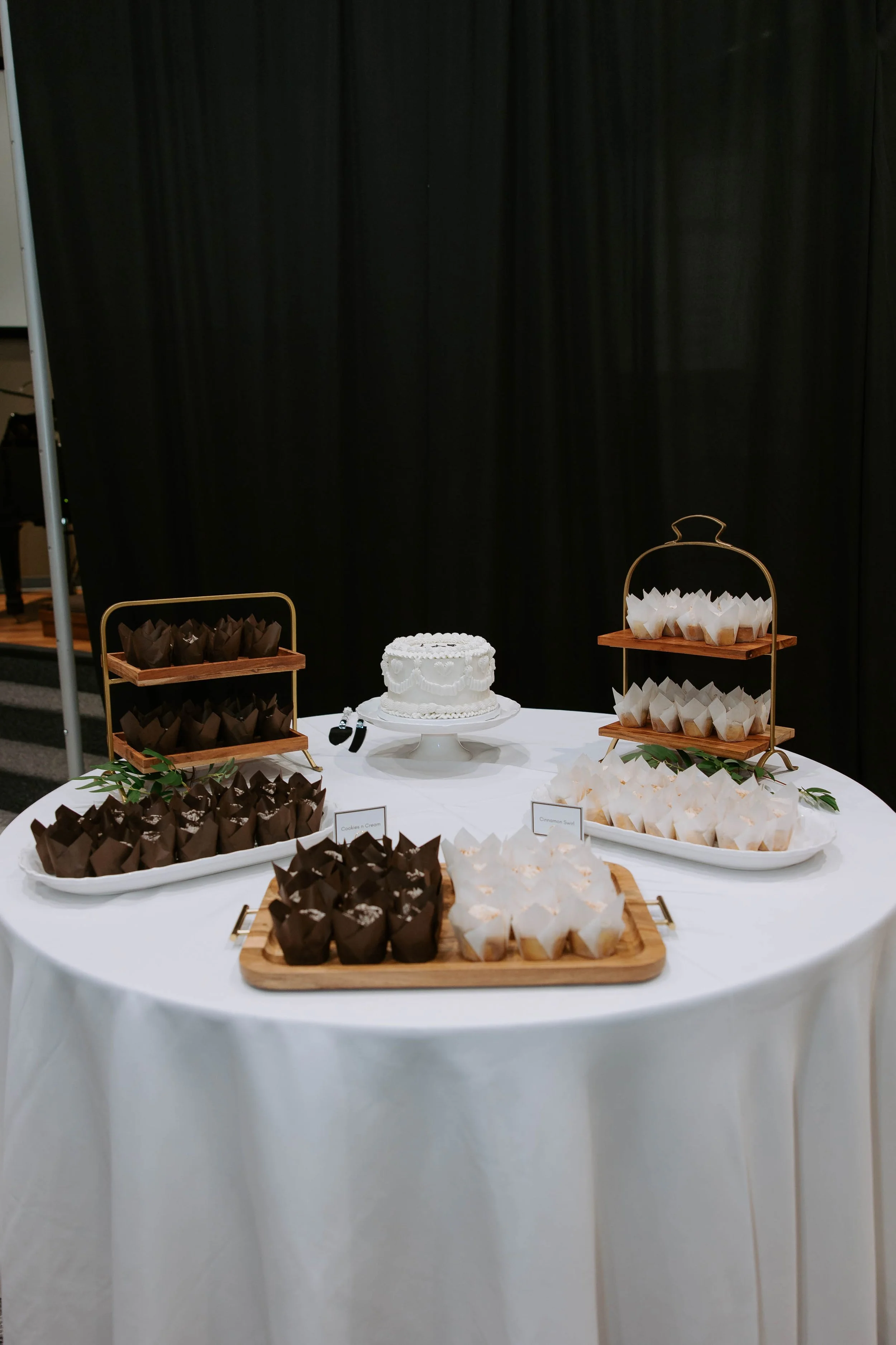 A table with a white tablecloth displaying a wedding cake and an assortment of cupcakes in chocolate and vanilla flavors, arranged on trays and stands, with greenery for decoration, against a black curtain backdrop.