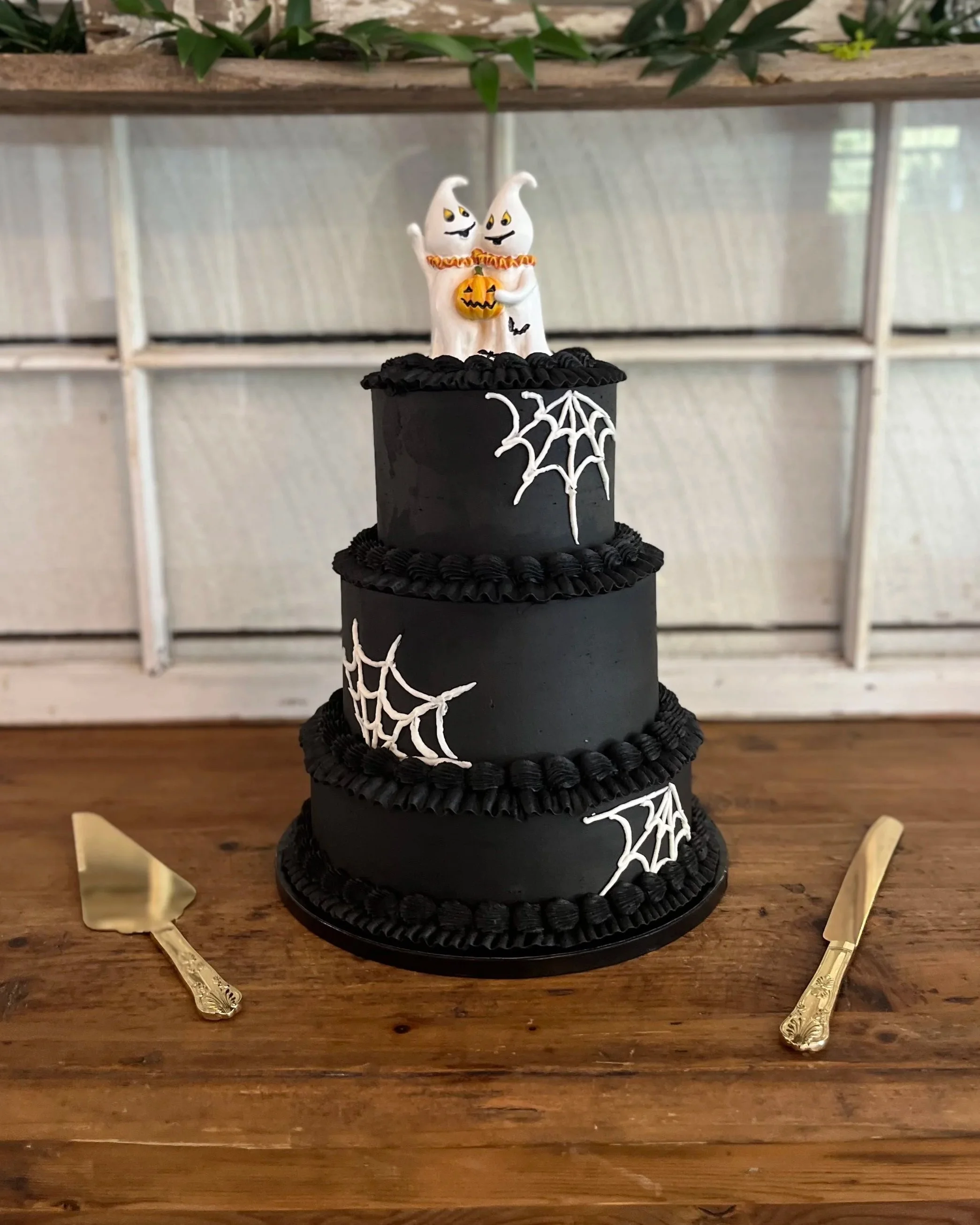 Three-tiered black Halloween cake with tiny ghosts on top, decorated with white spiderweb designs, set on a wooden table with cake knife and server.