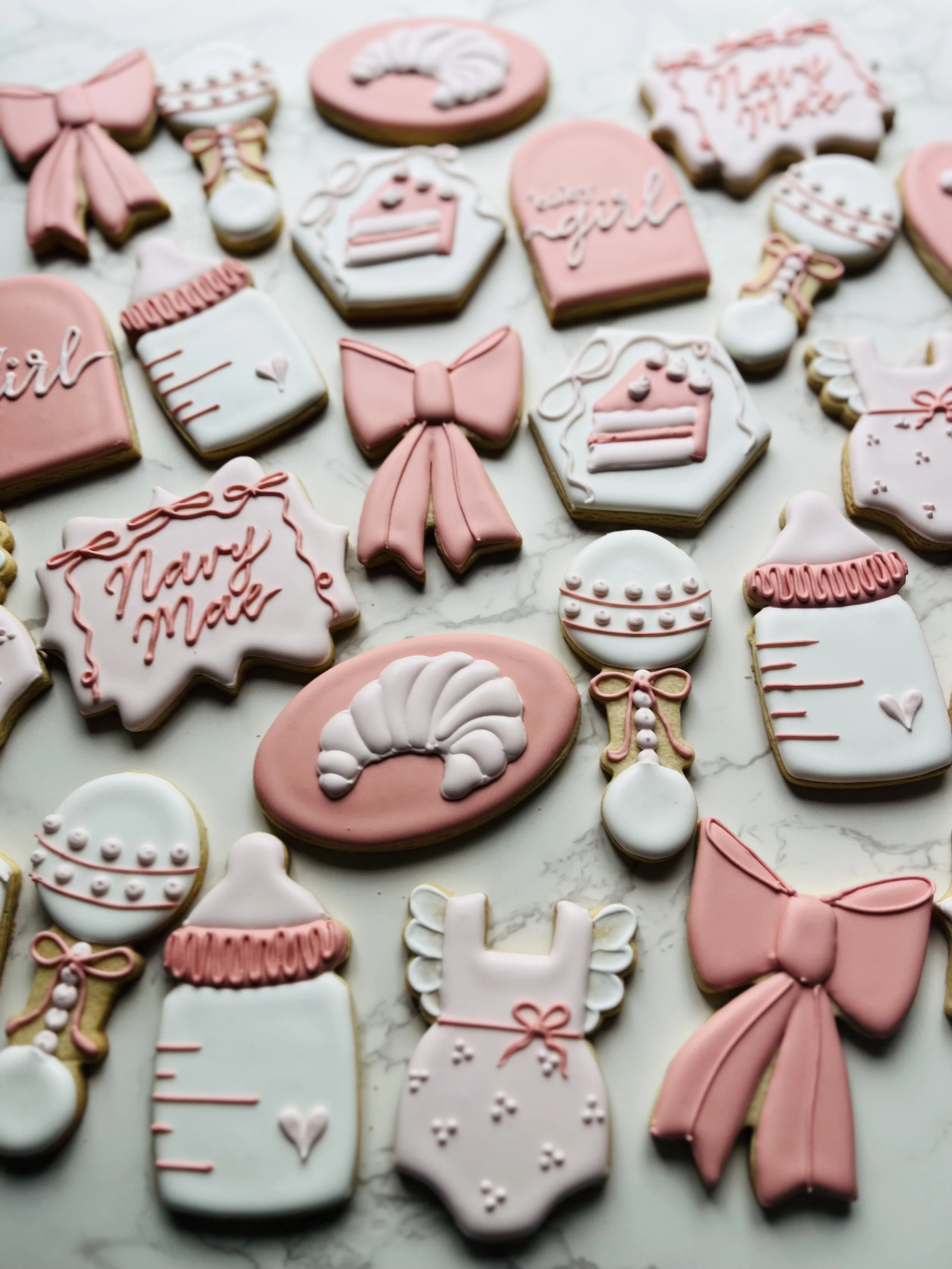 Assorted decorated cookies with pink, white, and beige icing, featuring bows, cake, gift box, and holiday themes, on a marble surface.