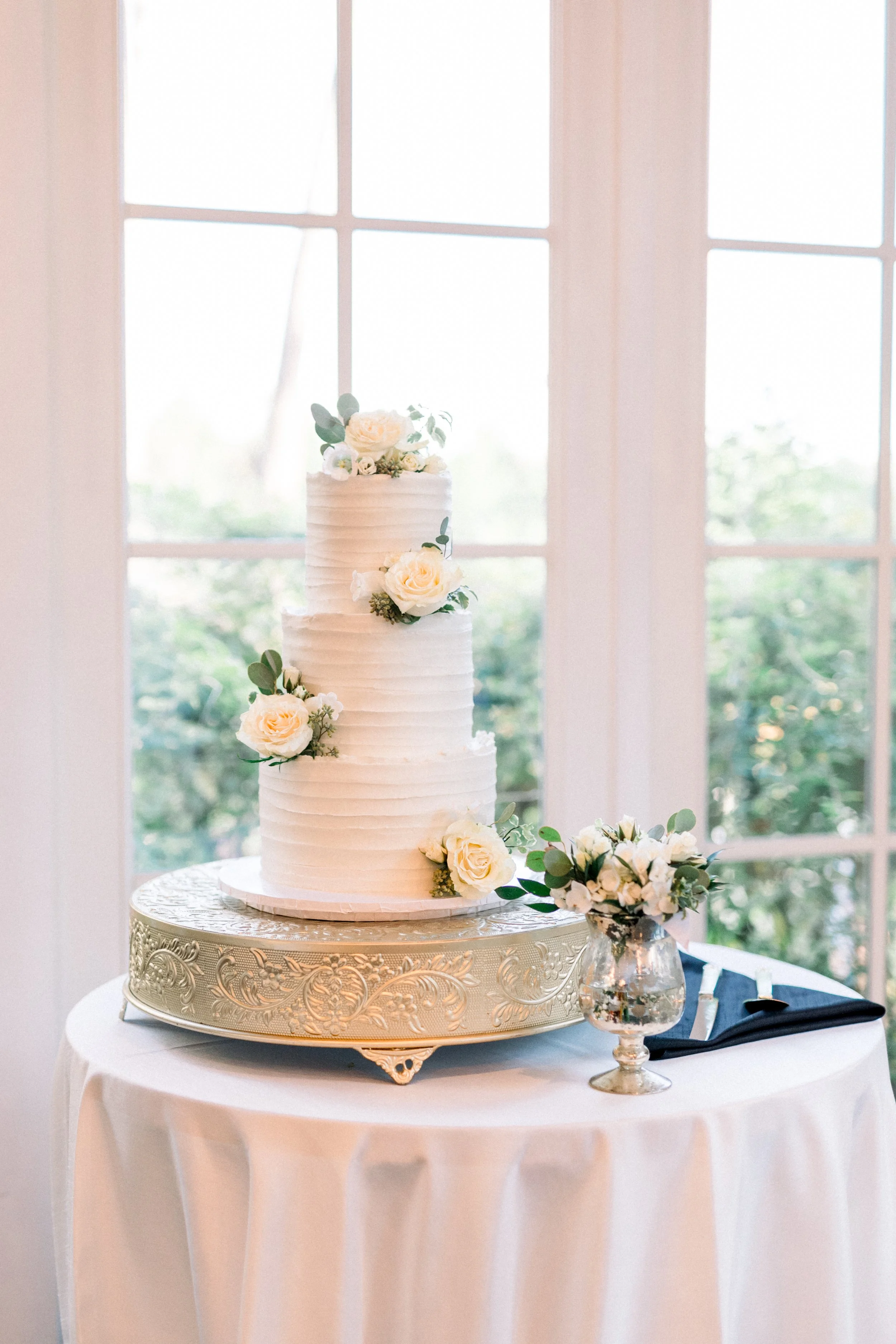 A tall white wedding cake decorated with white roses and greenery, placed on a silver ornate cake stand on a round table with a white tablecloth. There is a bouquet of white flowers and greenery in a glass vase beside the cake. The background shows large windows letting in natural light.