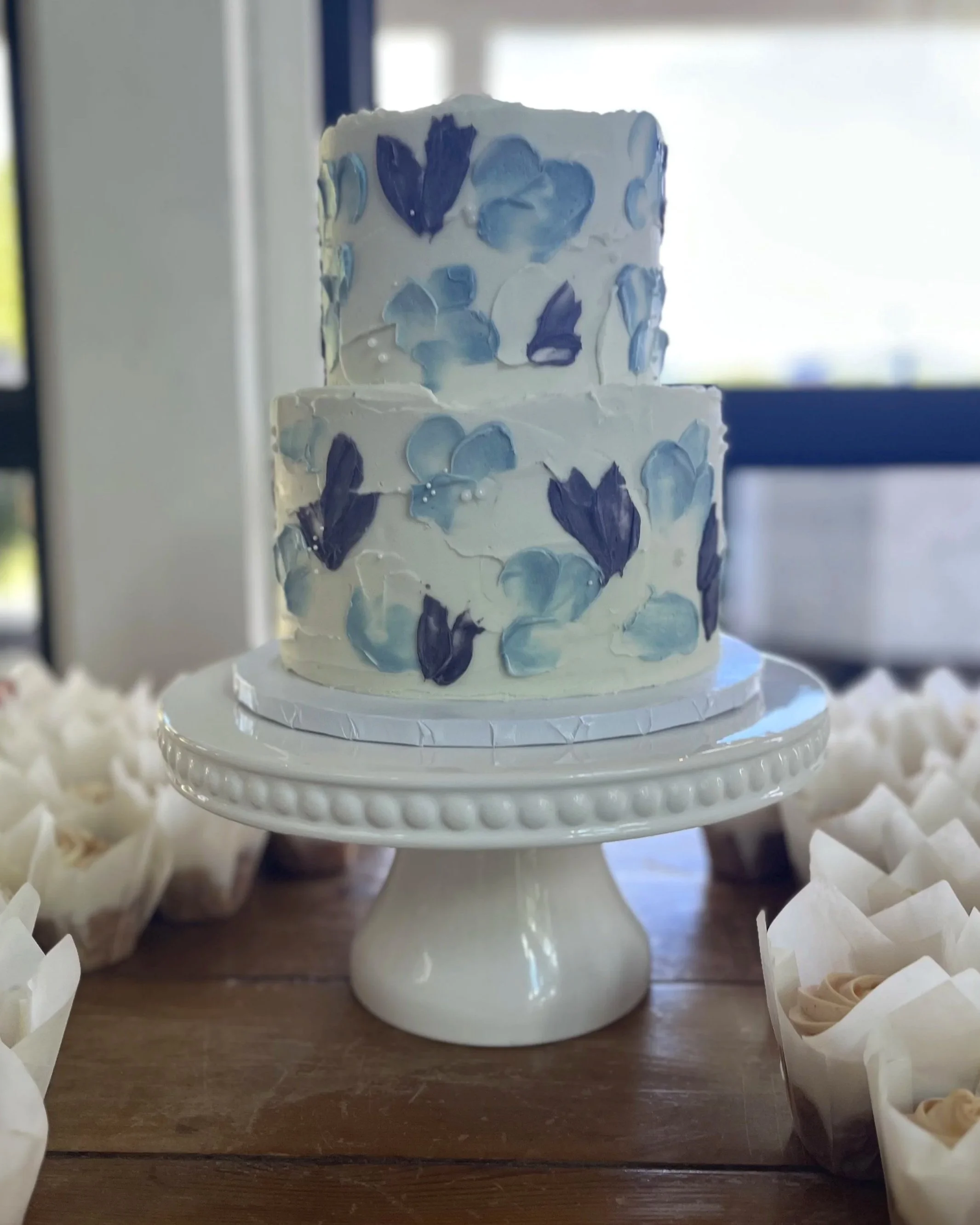 Two-tiered white wedding cake with blue and purple flower decorations on a white cake stand.