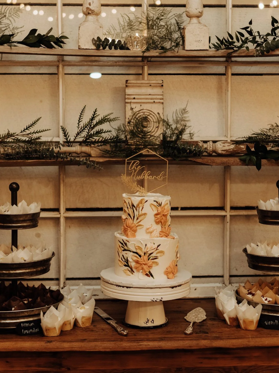 A tiered wedding cake decorated with peach and black floral designs, sitting on a wooden table with servers and desserts around it, and a decorative topper reading "The Hubbards".