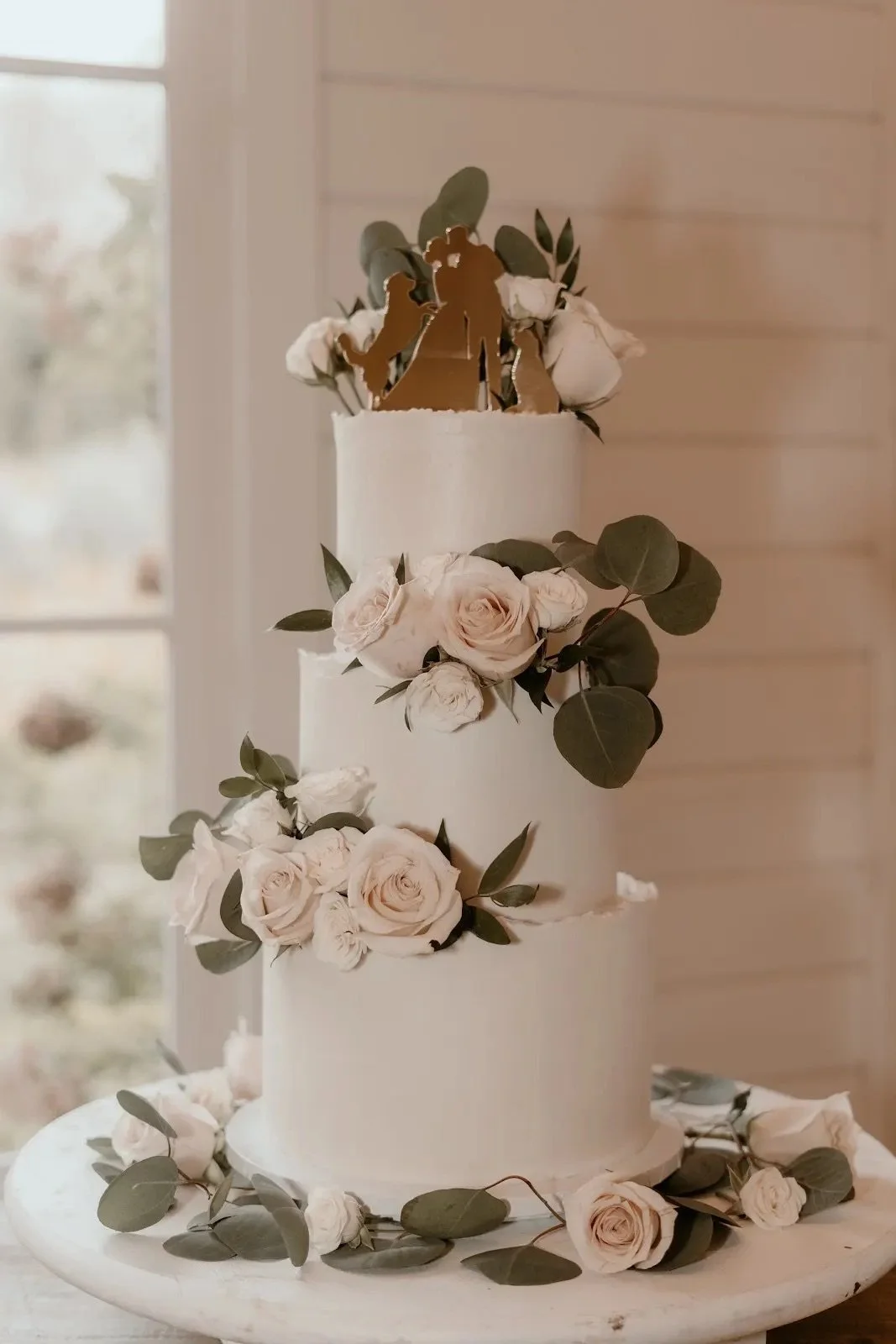 A three-tier white wedding cake decorated with white roses and green leaves, topped with a gold cake topper of a couple holding hands, set against a wooden wall background.