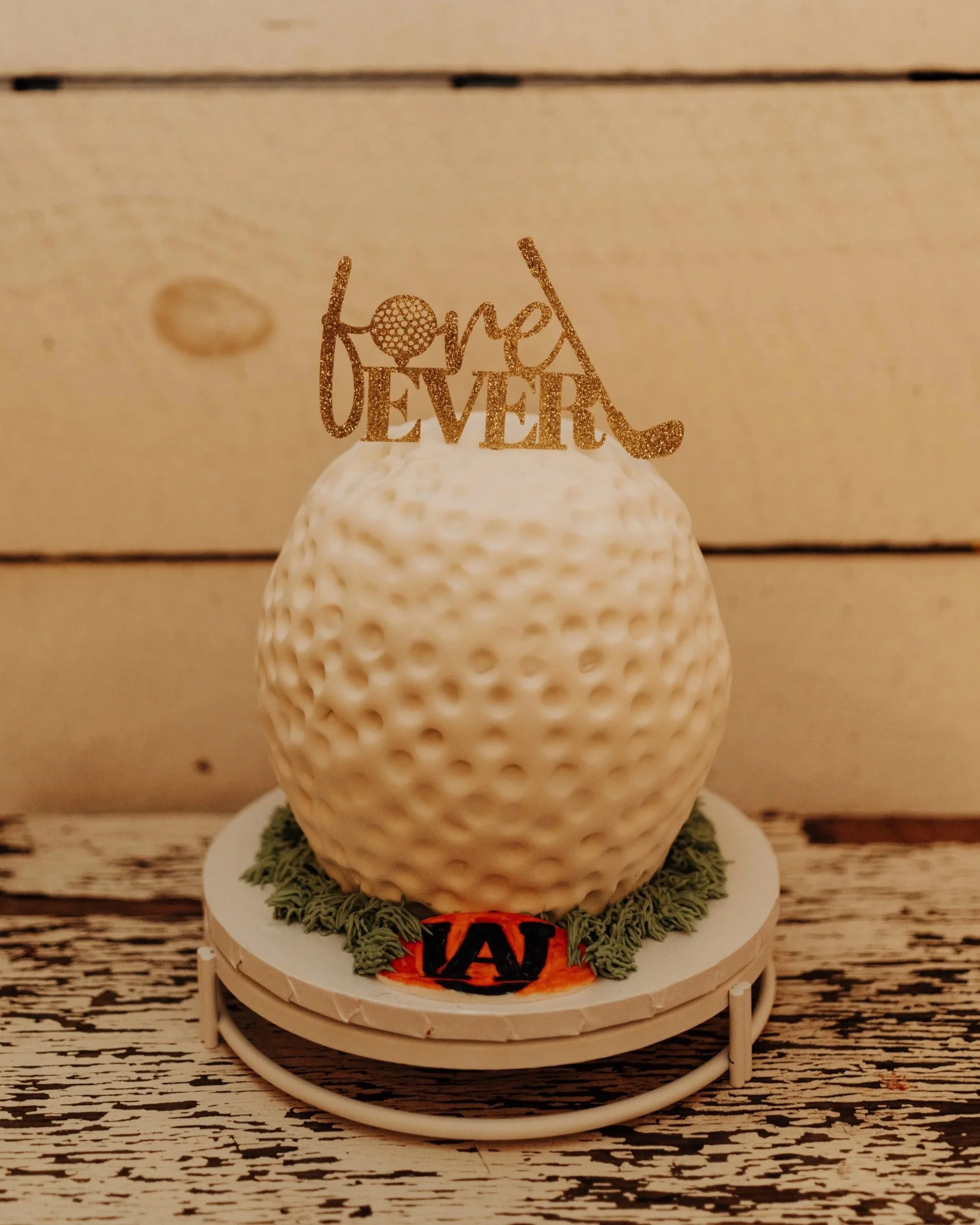 Decorative golf-themed cake with white textured frosting, a glittery gold topper that says 'fore EVER' with a golf ball replacing the 'o', and green frosting resembling grass around the cake on a white base.