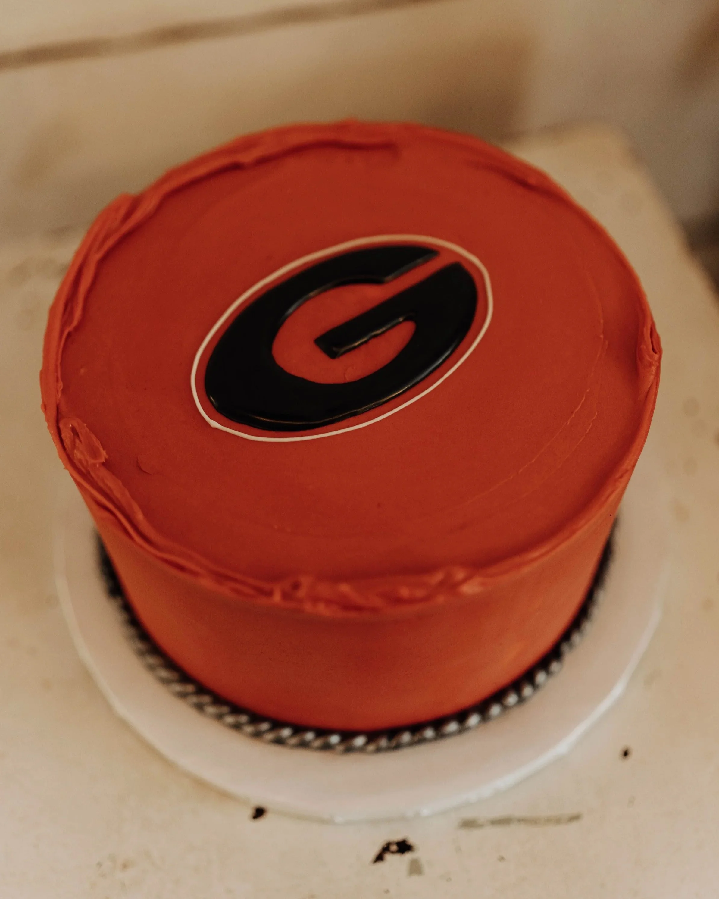 A round cake with orange frosting and a black and orange logo on top, sitting on a white surface.