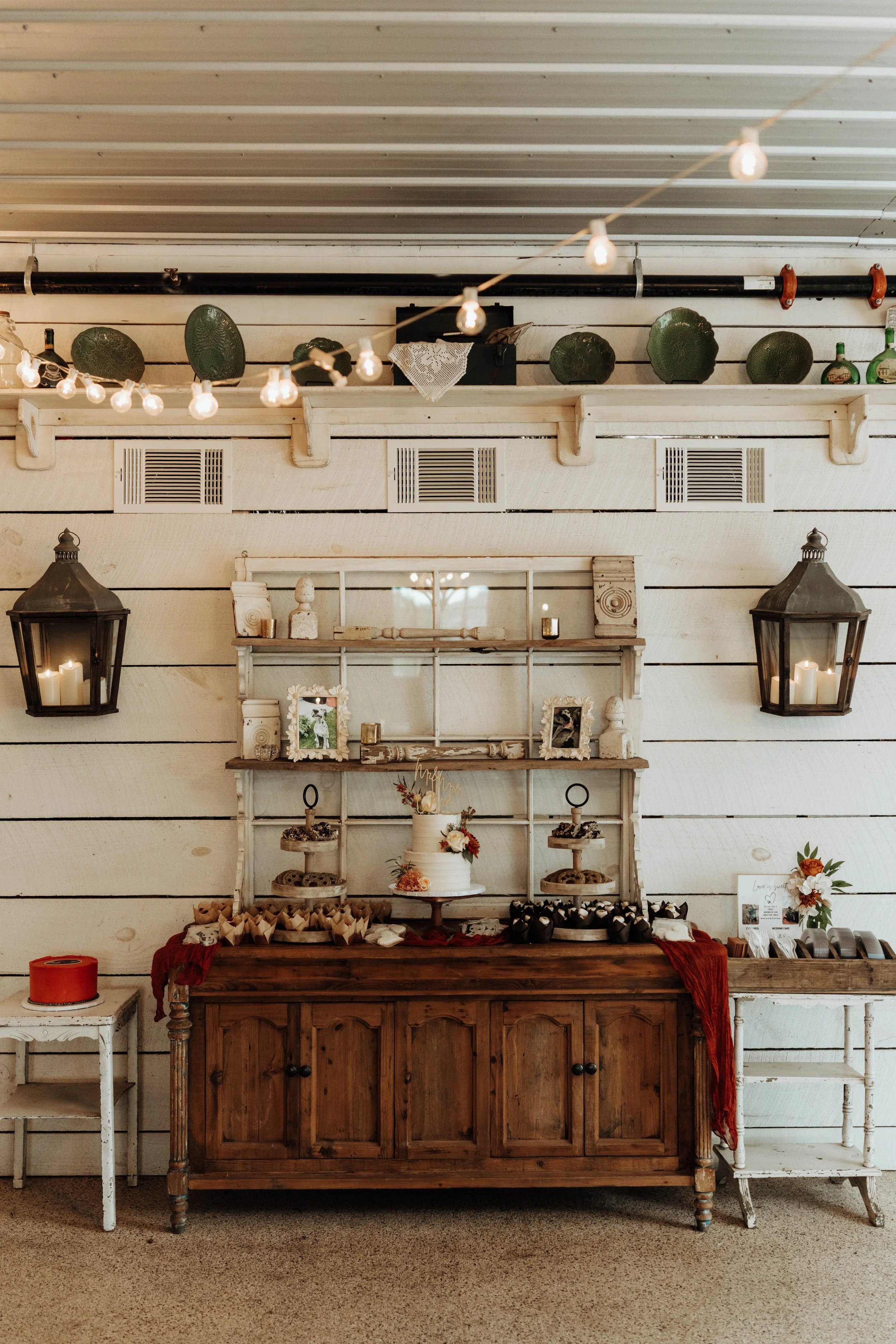 A dessert table setup with a cake and assorted treats, against a white shiplap wall, decorated with lanterns and string lights, in a rustic style venue.