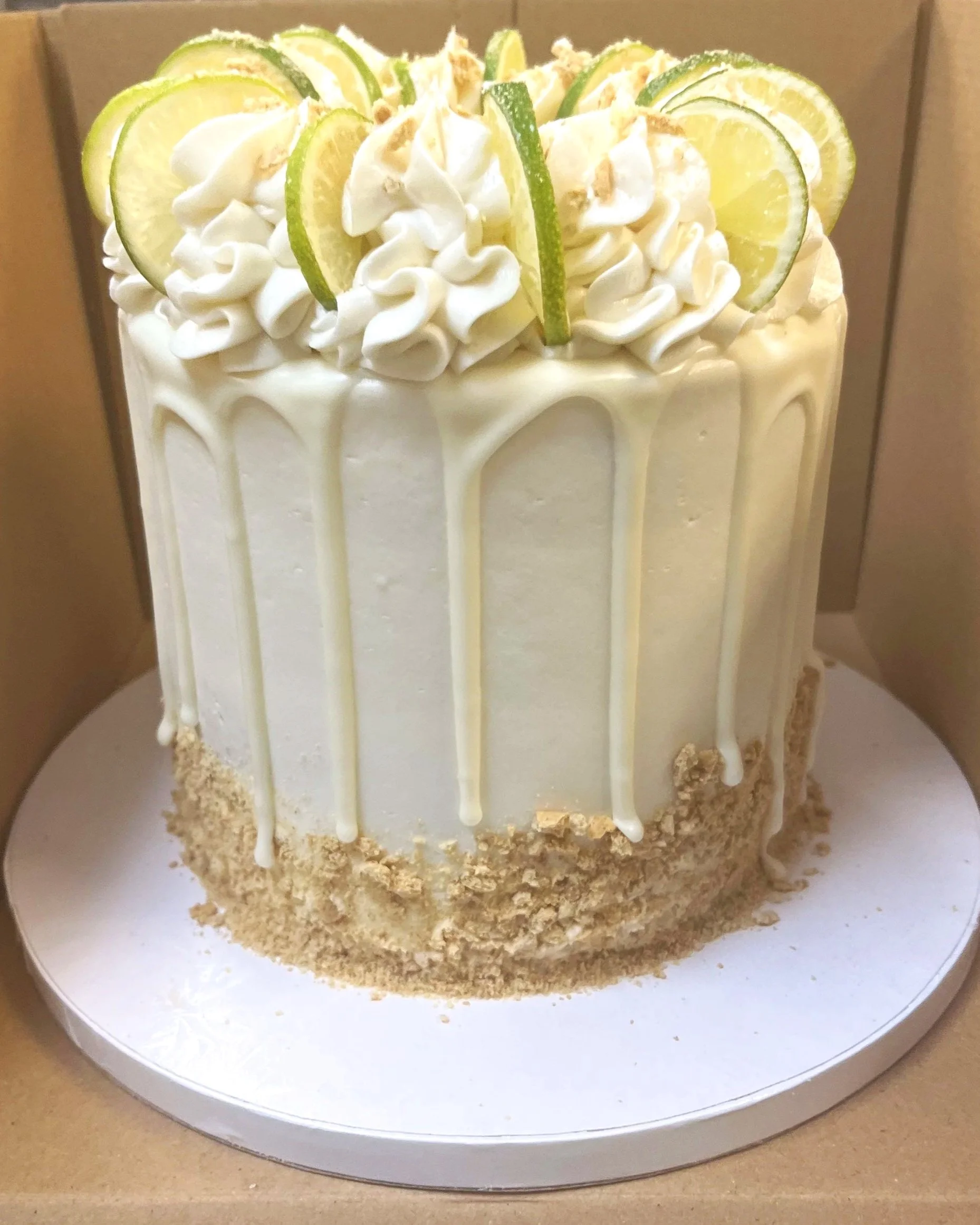 A tall white cake decorated with lime slices, white frosting, and white chocolate drips, with a crumbly base, placed on a white circular cake board.