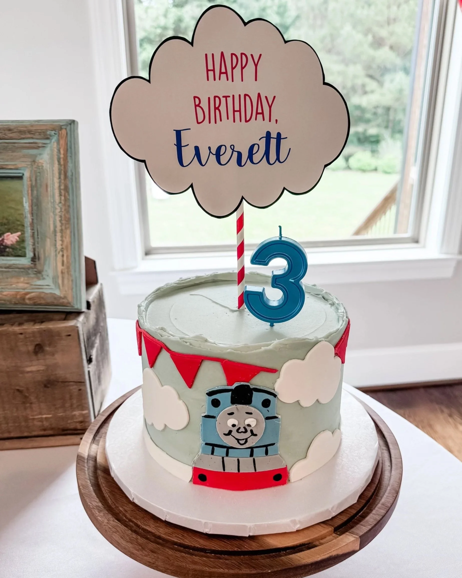 A Thomas the Tank Engine themed birthday cake with a number 3 candle, clouds, and a sign that says 'Happy Birthday, Everett' on top, placed on a wooden cake stand near a window.