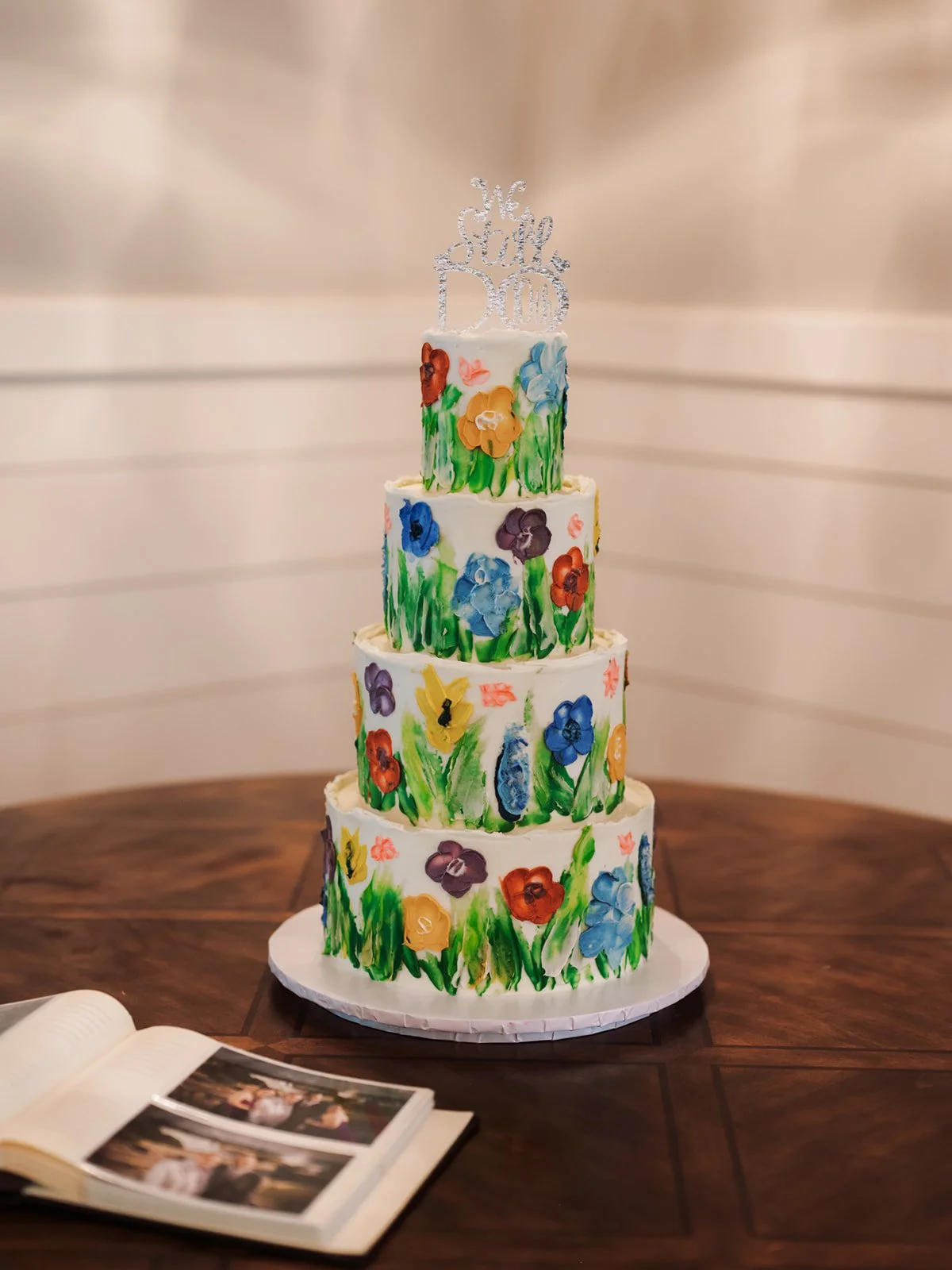 A four-tiered white wedding cake decorated with colorful hand-painted flowers and green grass-like borders. A silver topper reads "We Still Do." The cake is on a wooden table, with a photo album partially visible in the foreground.