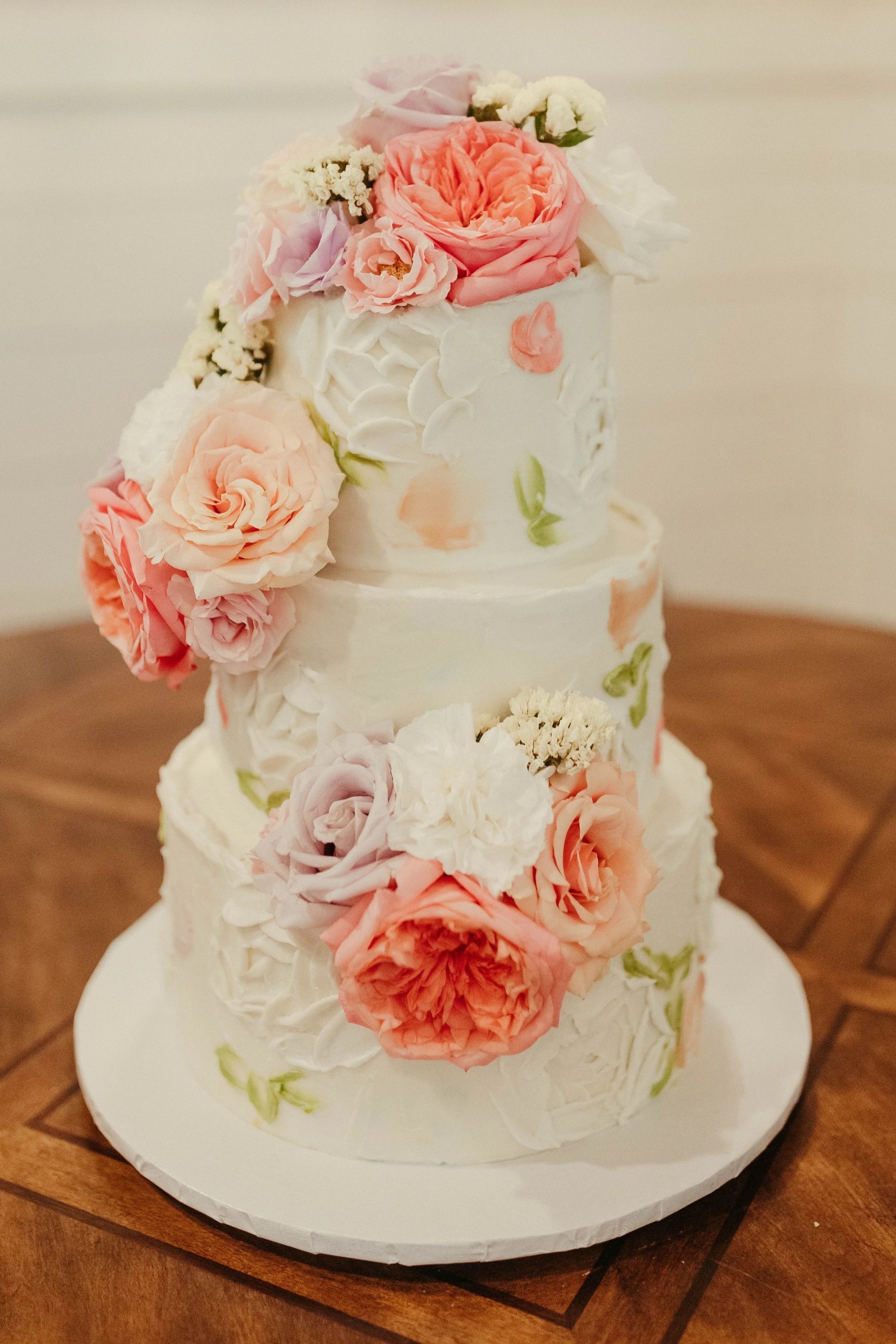 Three-tier white wedding cake decorated with pink and white roses and floral accents, sitting on a white cake board on a wooden table.