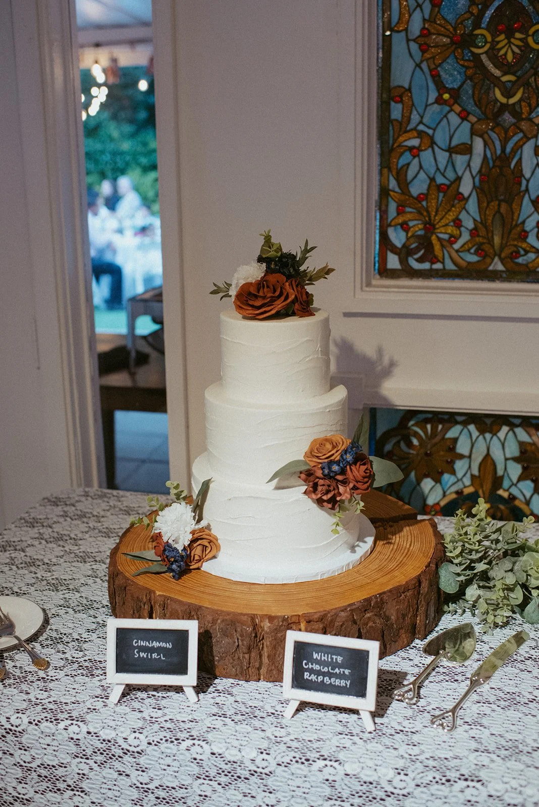 A three-tiered white wedding cake decorated with brown, orange, and blue flowers, sitting on a wooden slab on a lace tablecloth. Small chalkboard signs label the flavors: 'Cinnamon Swirl' and 'White Chocolate Raspberry'.
