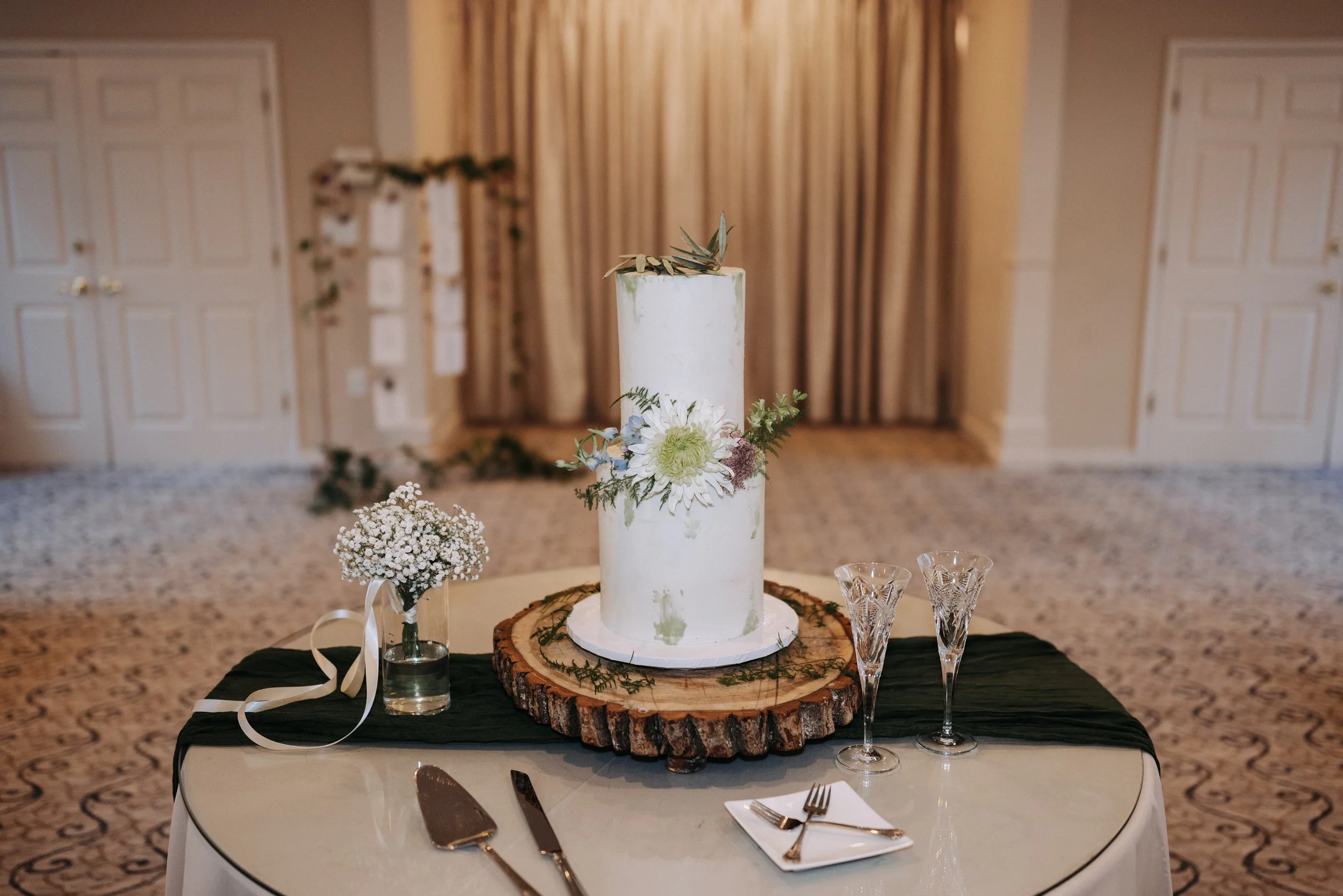 A two-tier white wedding cake decorated with white and green flowers, placed on a round wooden slab on a black and white table cloth. There are two empty crystal champagne glasses, a small bouquet of white baby's breath, and cake serving utensils on the table.