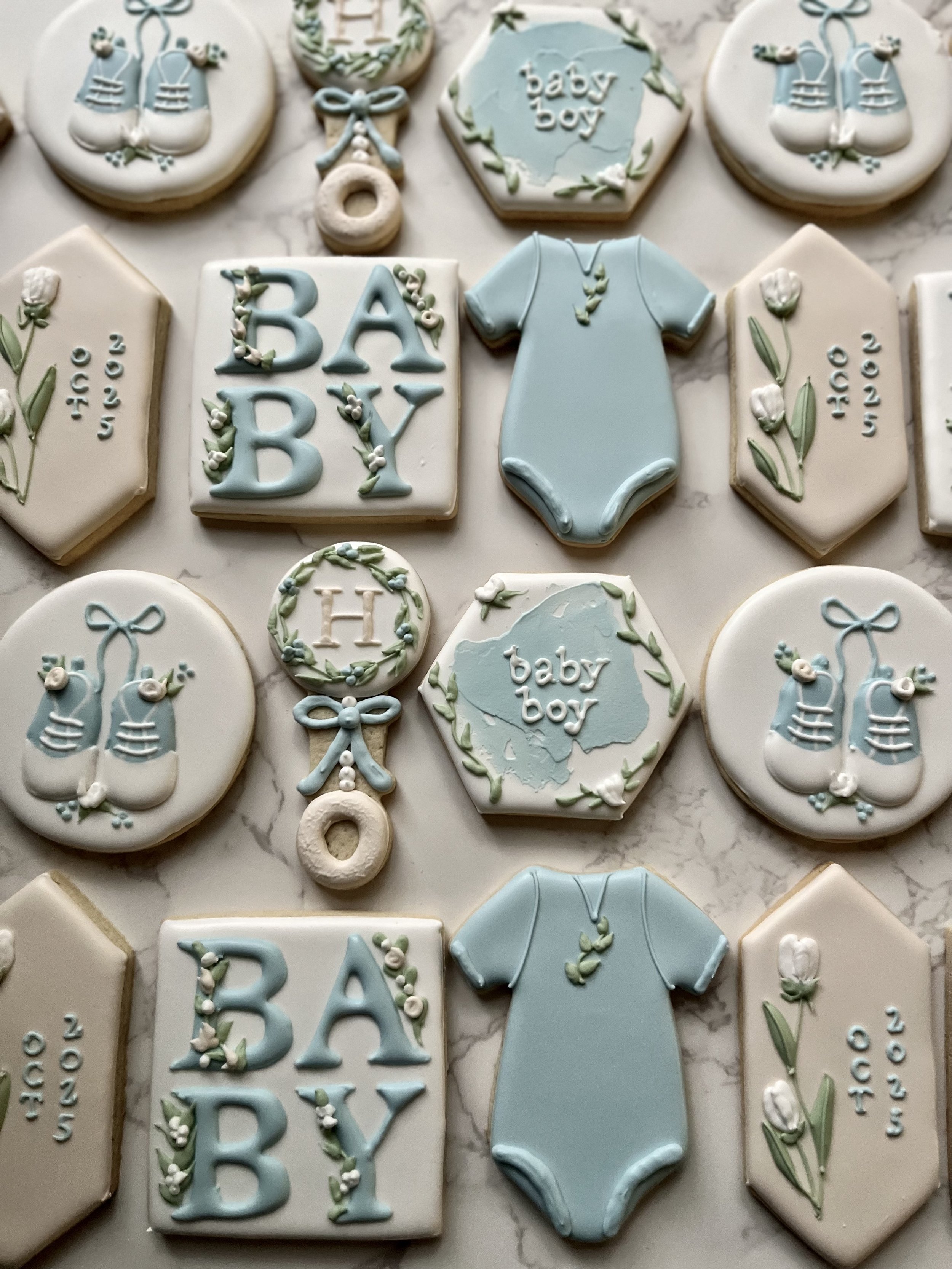 Collection of decorated cookies with baby and maternity themes, featuring baby onesies, the word 'baby boy', flowers, booties, and the date October 2022, in pastel blue, white, and green colors.