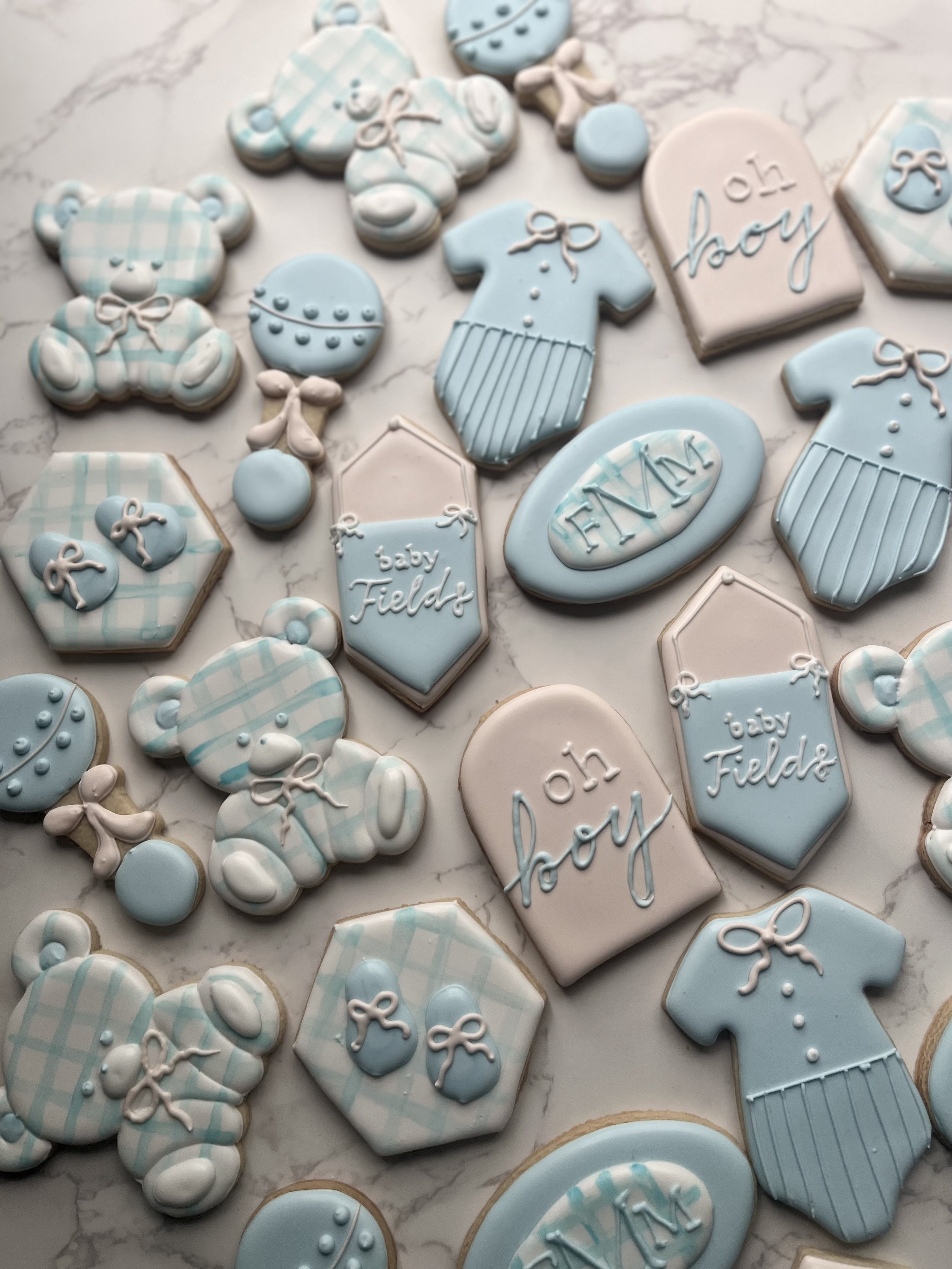 Assorted baby shower cookies decorated in pastel blue and white, featuring teddy bears, pacifiers, baby bottles, onesies, and plaques with phrases like 'oh boy,' 'baby Fields,' and 'FAM.'