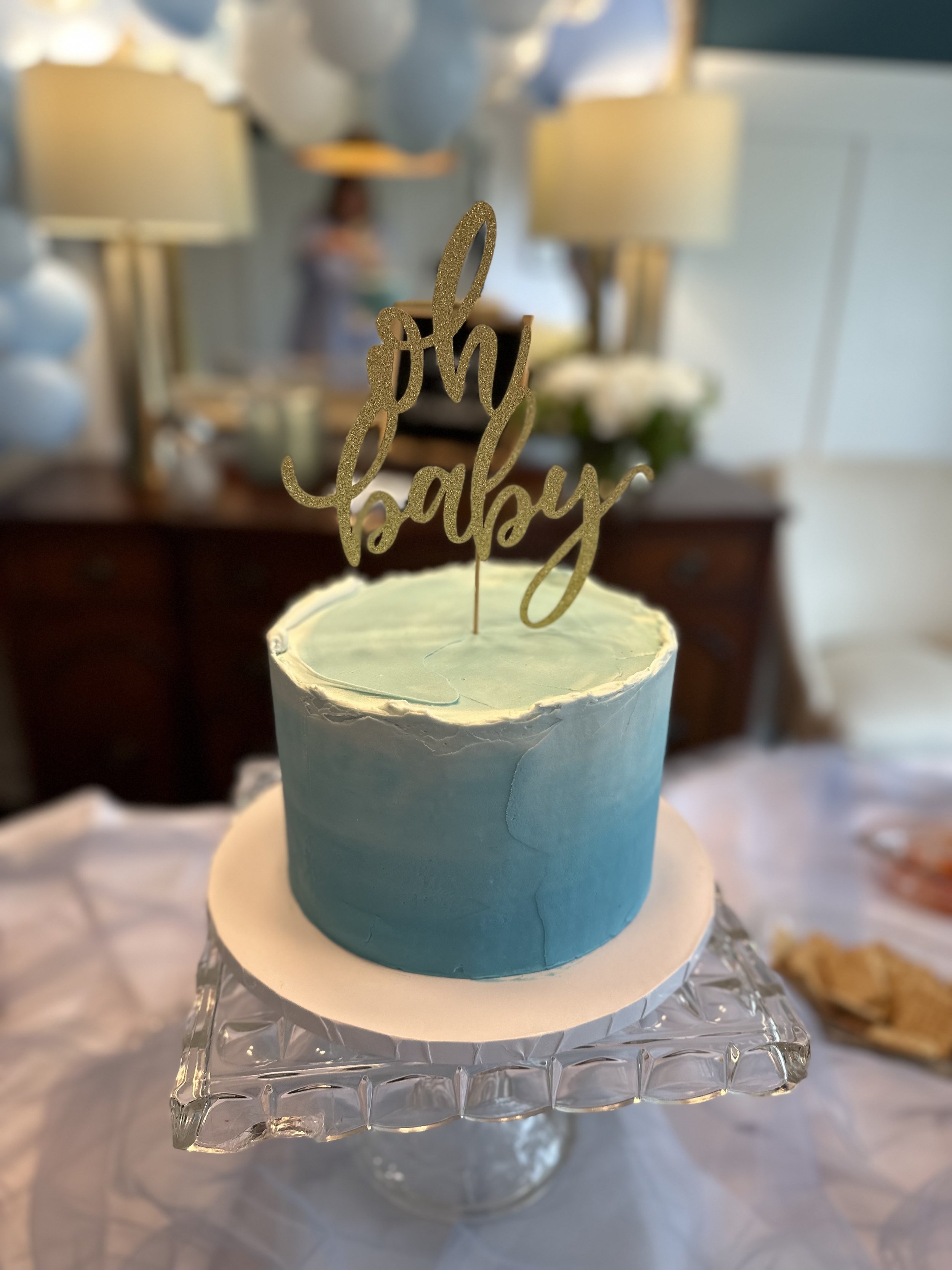 A cake with blue and white frosting on a glass stand, topped with a gold glittery "oh baby" cake topper, at a celebration setting.