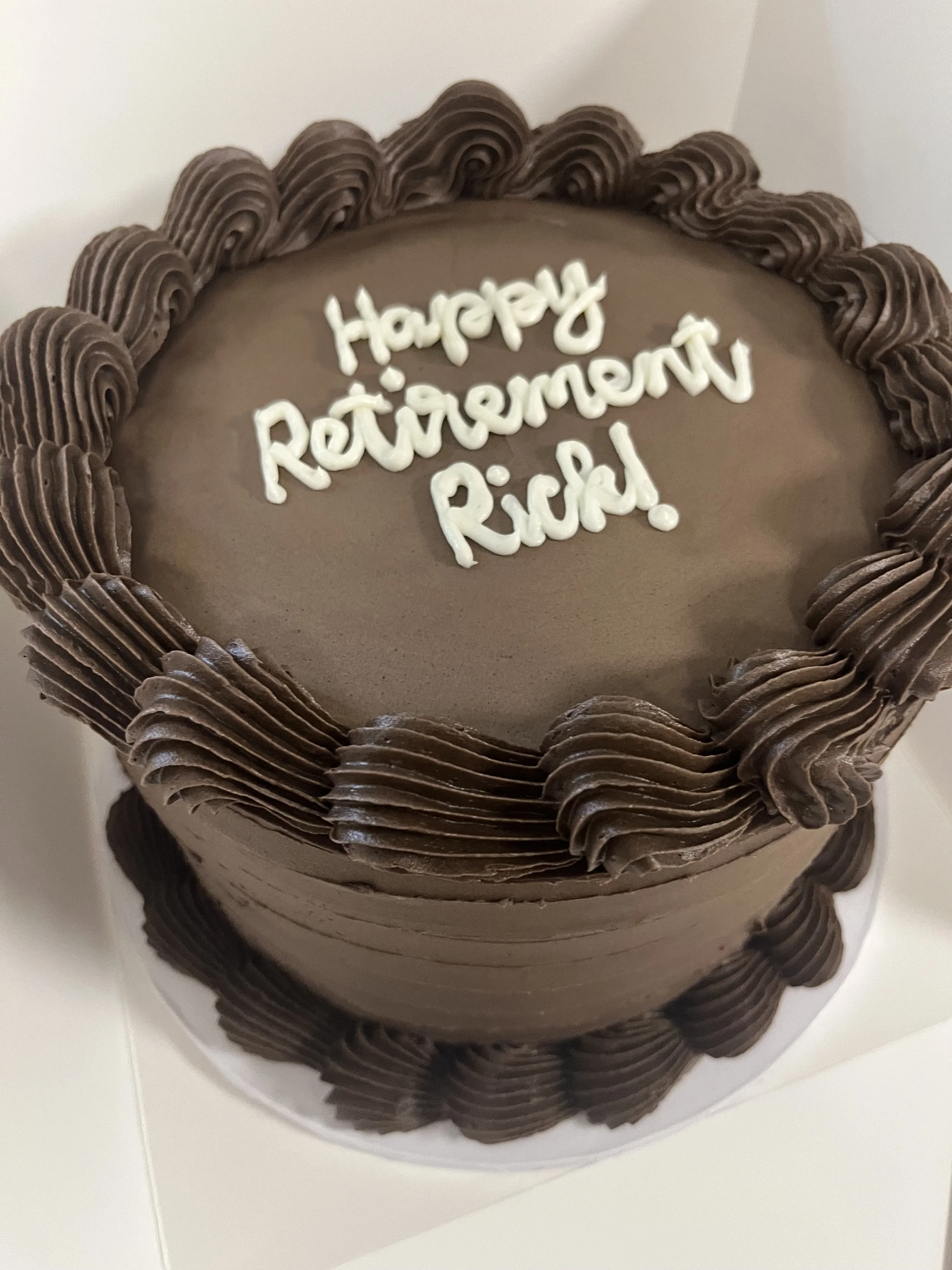 Chocolate birthday cake with frosting border and white icing message that reads 'Happy Retirement Ricky'.