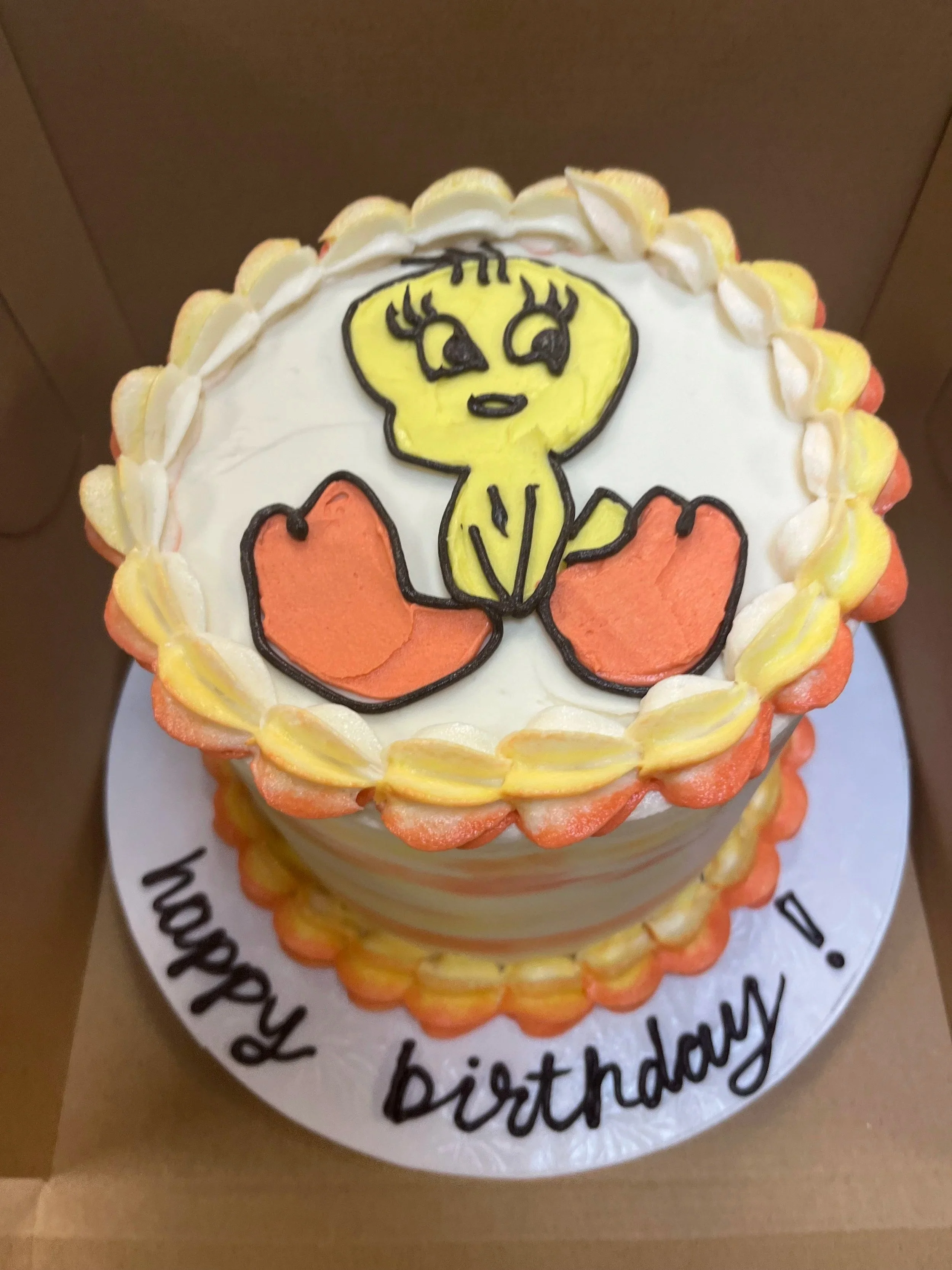 A birthday cake decorated with a cartoon character drawing of a yellow chick sitting on orange eggs, with a yellow and orange piped border."