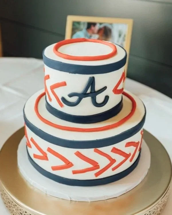 Two-tier cake with white icing, navy blue and red decorations, and a letter 'A' on top. The cake has red arrow patterns and is placed on a gold-colored board. In the background, there's a blurred photo frame with a picture of a couple.