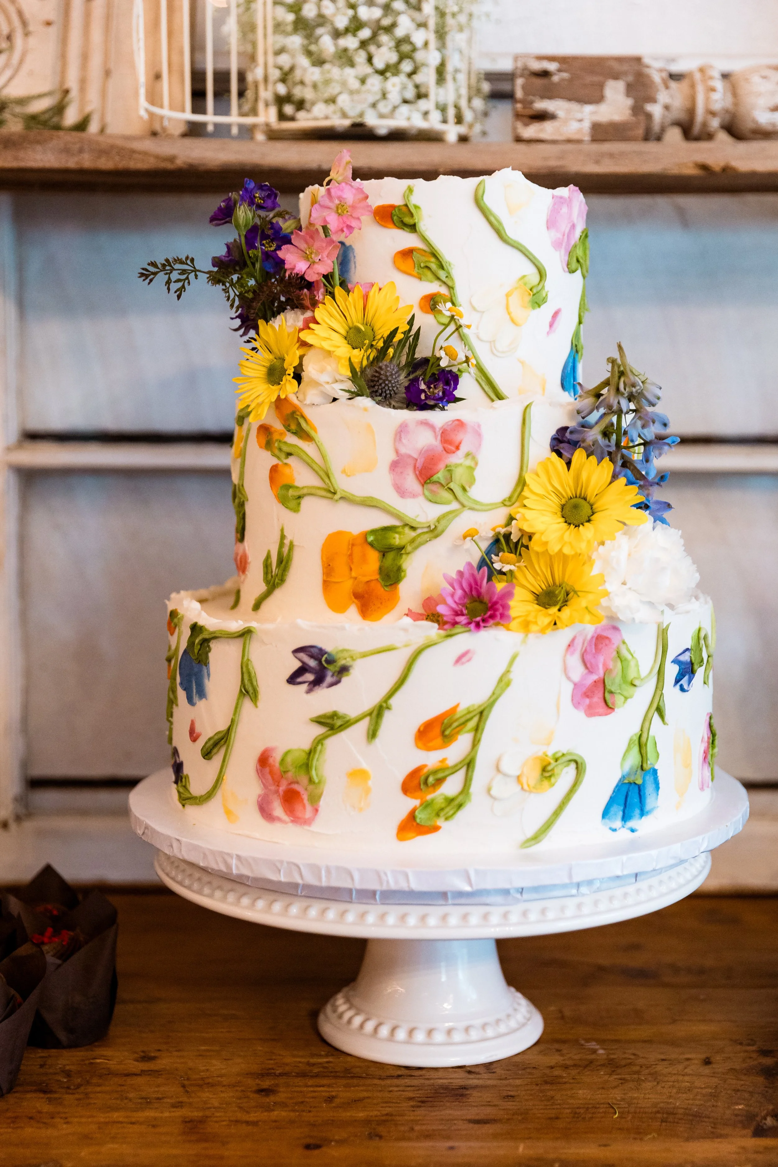 Three-tier white wedding cake decorated with colorful hand-painted floral designs and fresh flowers, placed on a white cake stand on a wooden table.