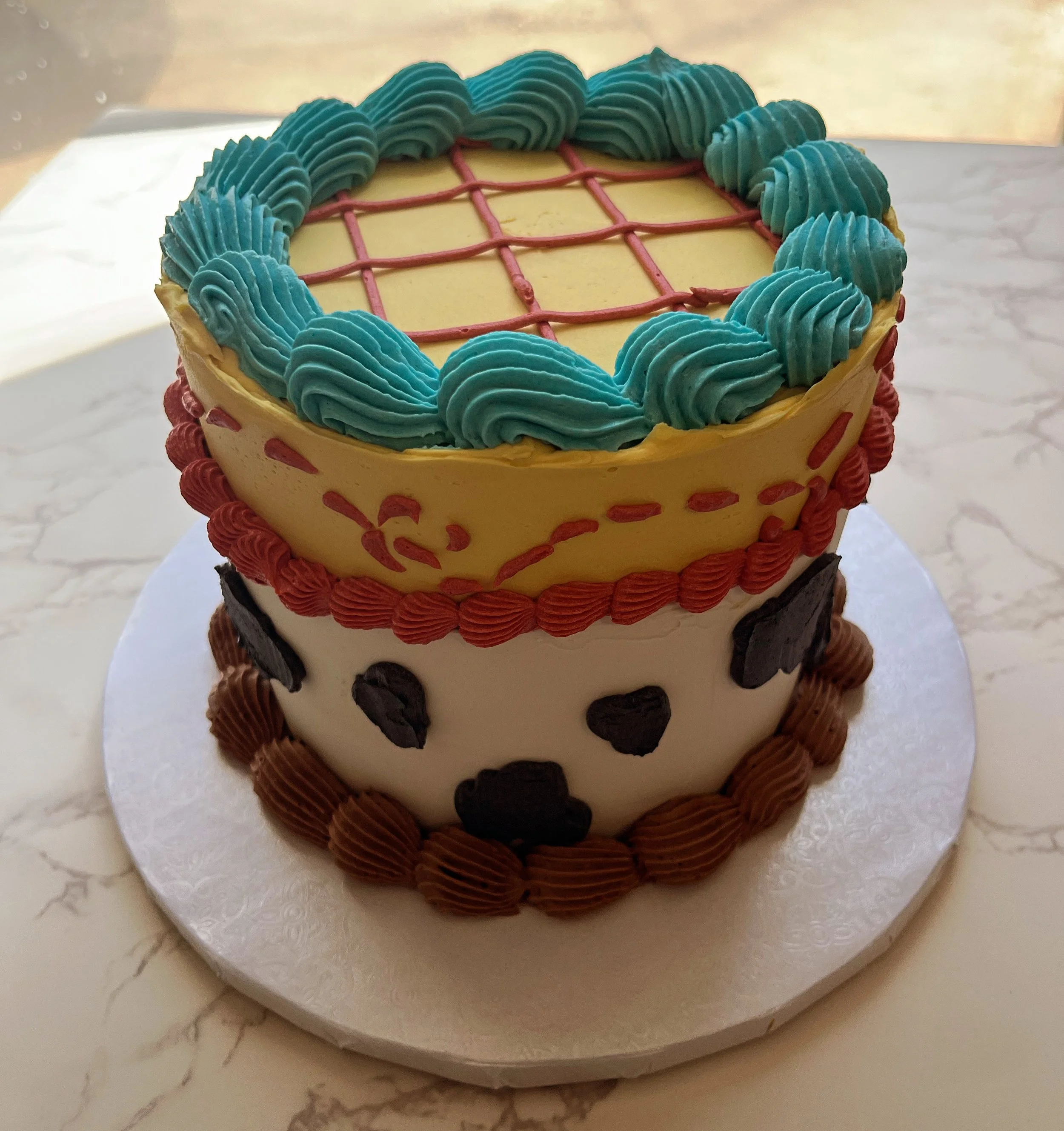 A two-tiered cake decorated with blue, red, black, and brown icing. The top tier has a yellow icing surface with red lines forming a grid and blue swirls around the edge. The bottom tier features white icing with black spots and a brown rosette border at the base.