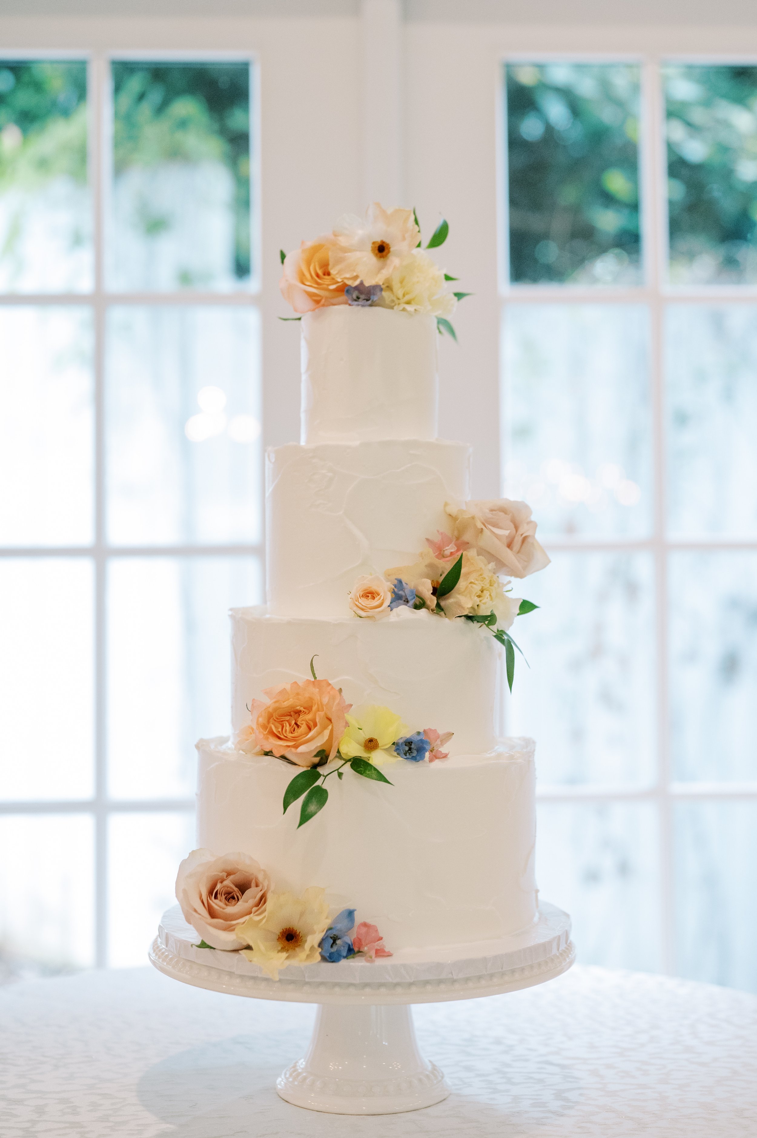 A white, four-tier wedding cake decorated with pastel-colored roses, peonies, and greenery, placed on a white cake stand in front of a window.