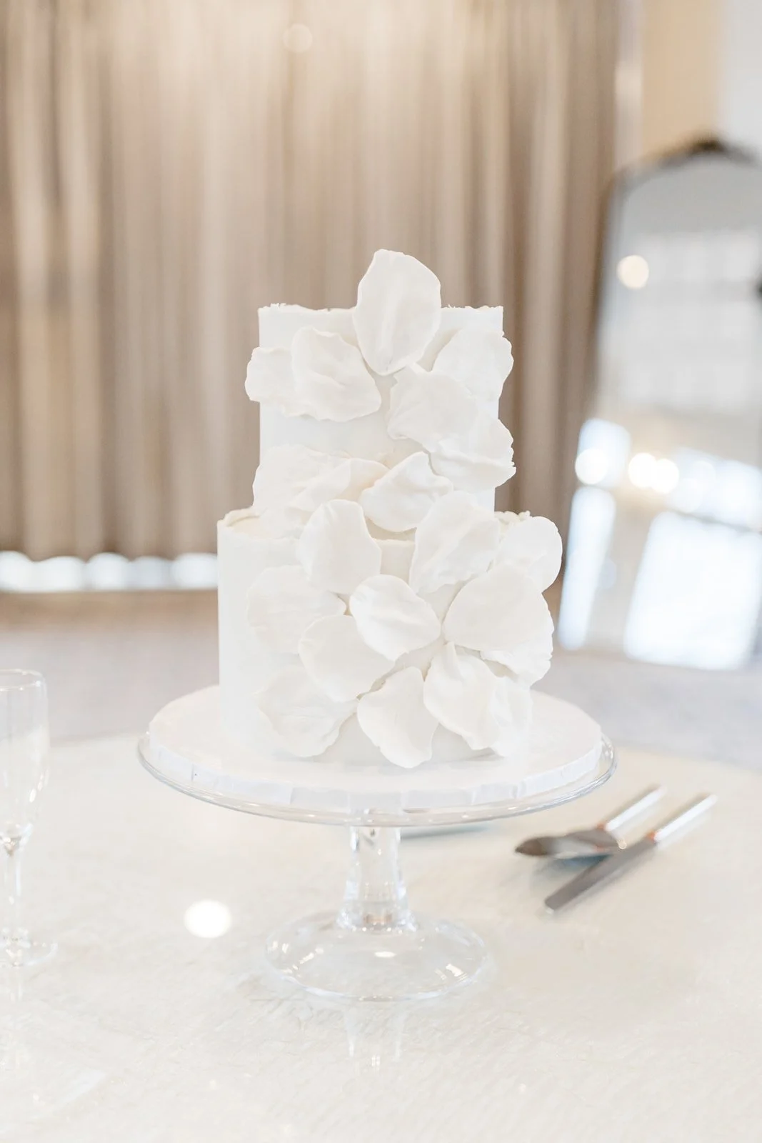 A white, multi-tiered cake decorated with white petal-like sugar flowers on a glass cake stand, set on a table with a white tablecloth, silver utensils nearby, and a blurred background with beige curtains.