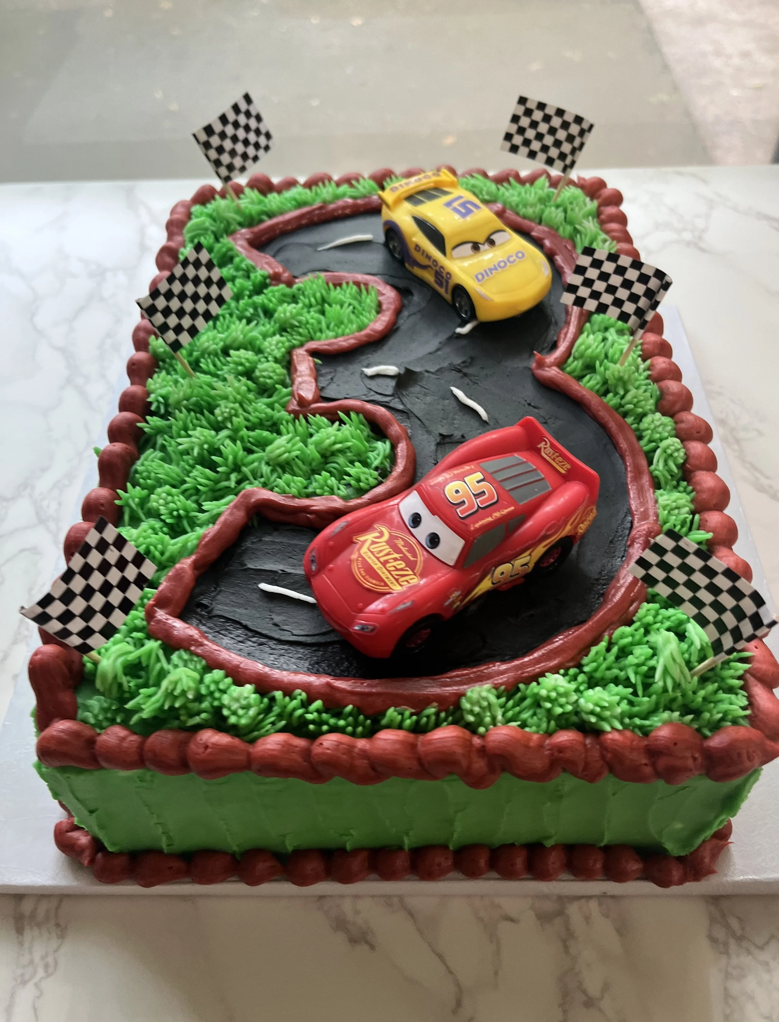 A rectangular birthday cake decorated with a racing theme, featuring green icing grass, a black road with red icing borders, and two toy cars resembling Lightning McQueen and another race car, with five checkered flags.