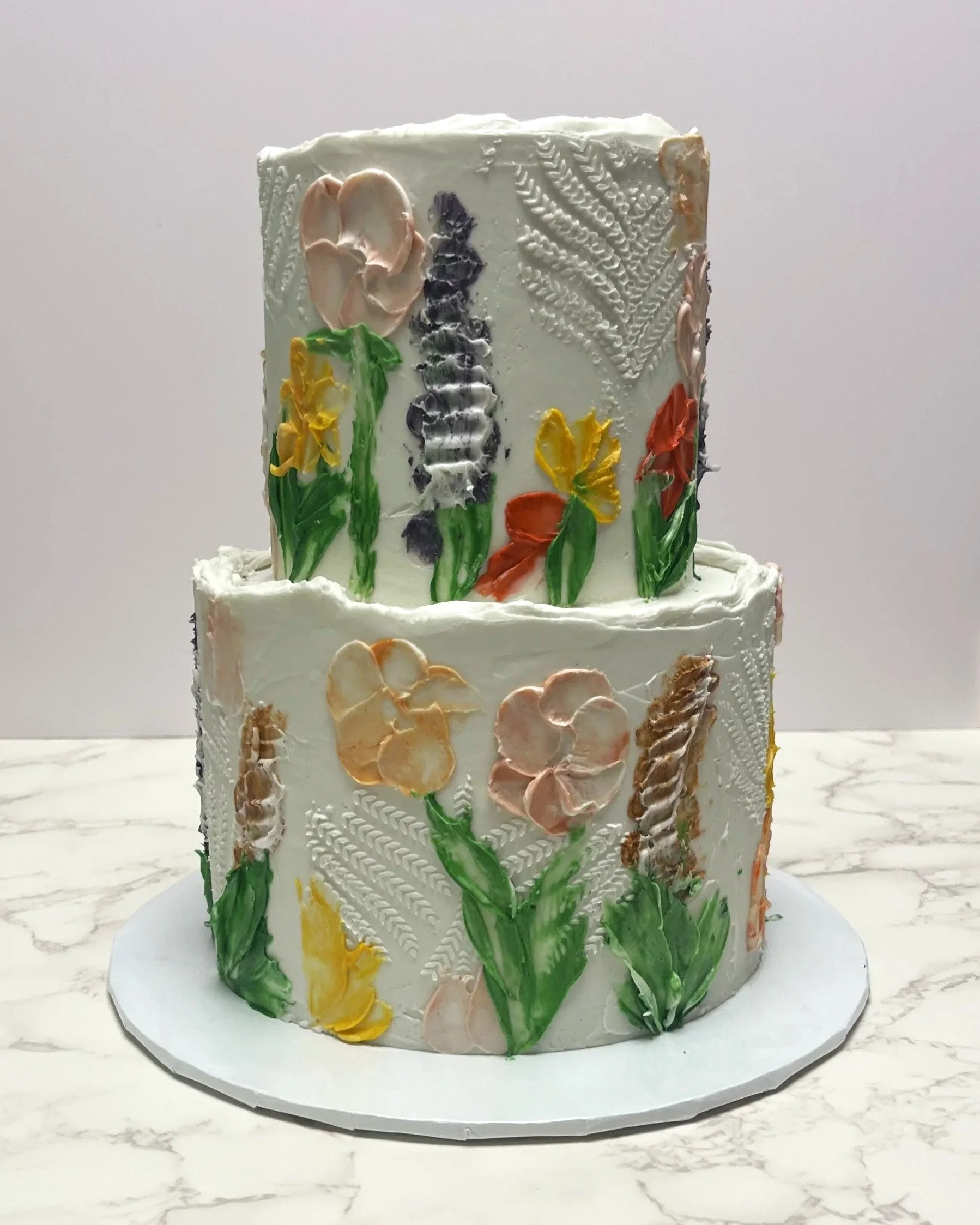 Two-tiered white cake decorated with colorful buttercream flowers and textured piping designs with a plain white background.