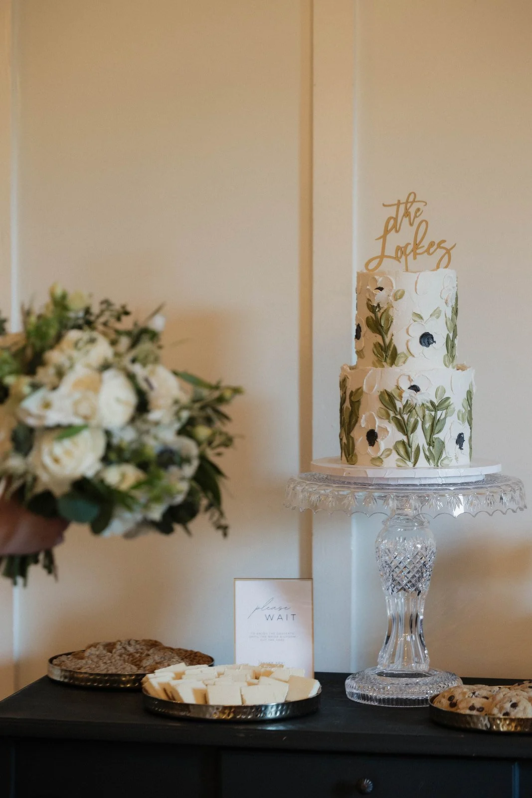 A two-tiered white wedding cake decorated with green leaves and dark berries on a glass pedestal, with a gold topper reading 'the Lopes'. A bouquet of white flowers is blurred in the foreground, and cookies and cheese are on trays on a black table.
