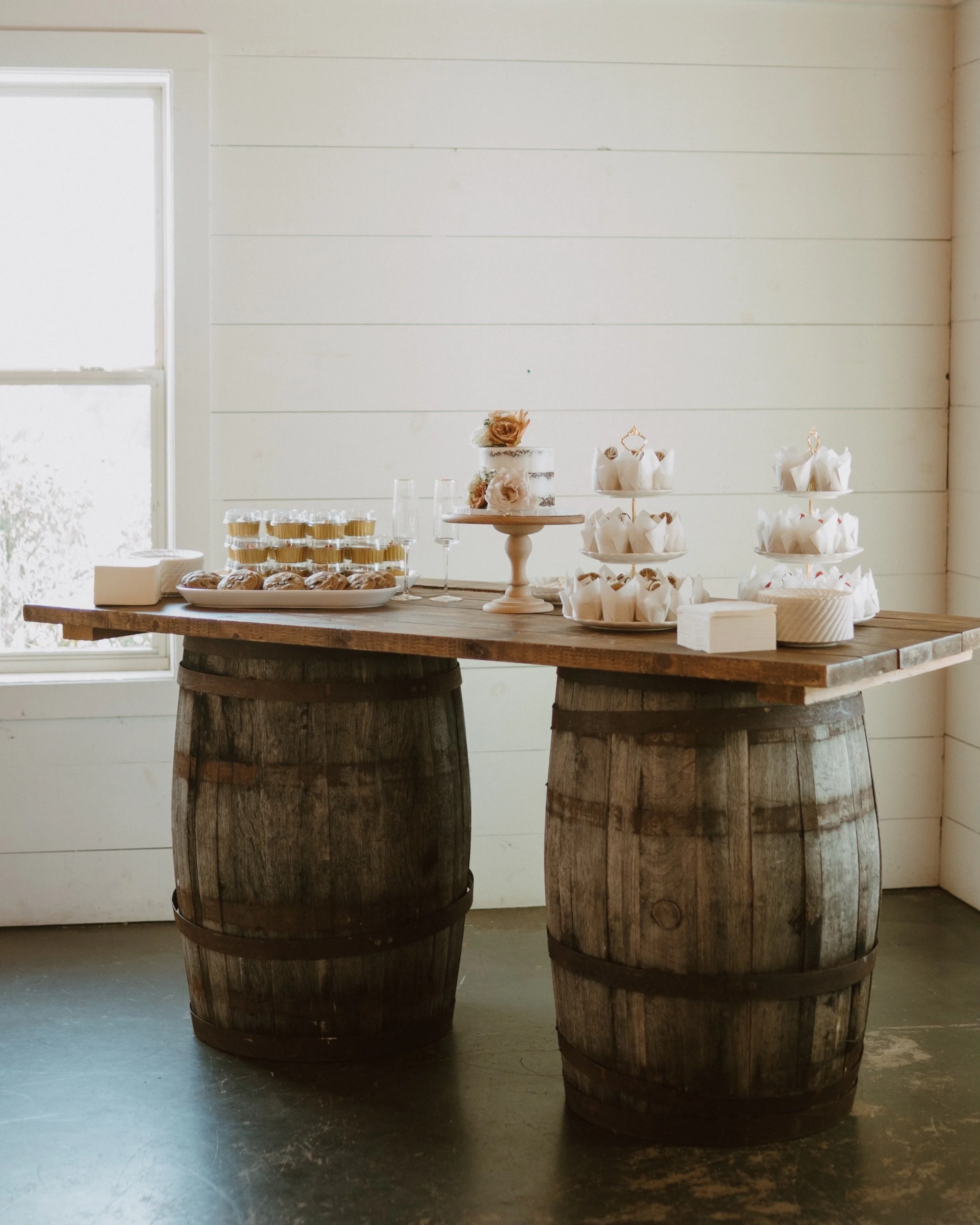 Decorative table setup with a tiered tray of cupcakes, a cake with flowers, and glasses of champagne on a rustic wooden table made from barrels, in a room with white paneled walls and a window.