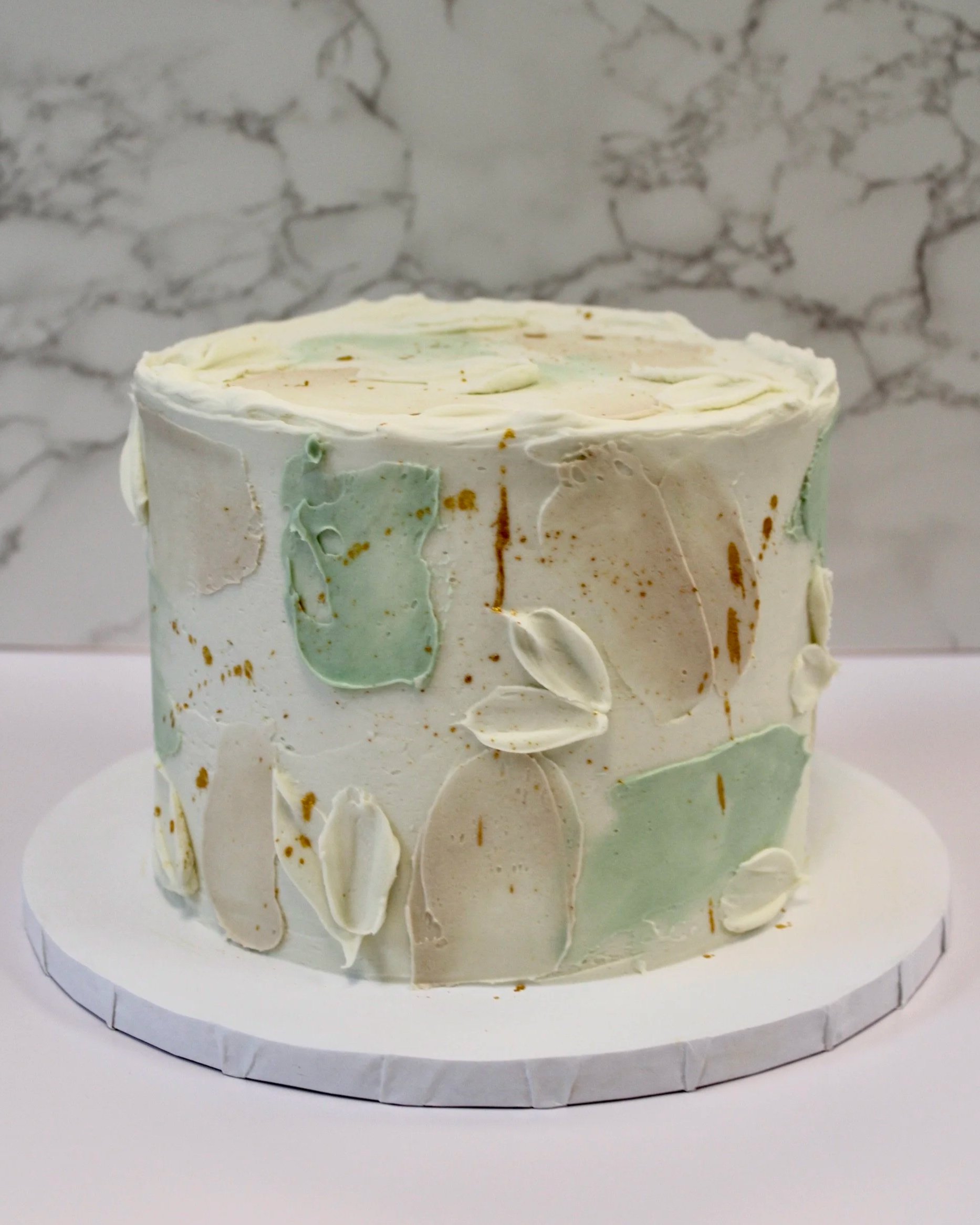 A round white cake with pastel green and white decorative icing on a white cake board against a marble background.