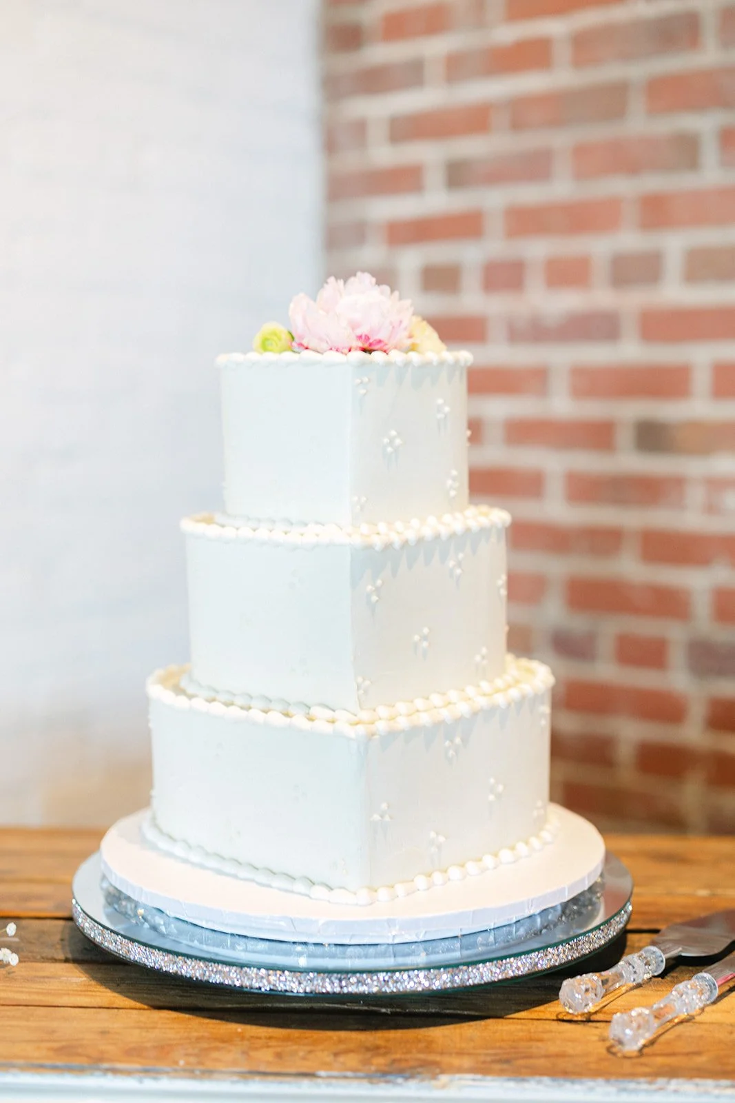 A three-tiered white wedding cake decorated with small edible pearls and flowers on top, placed on a wooden table with cake serving utensils nearby, against a brick wall background.