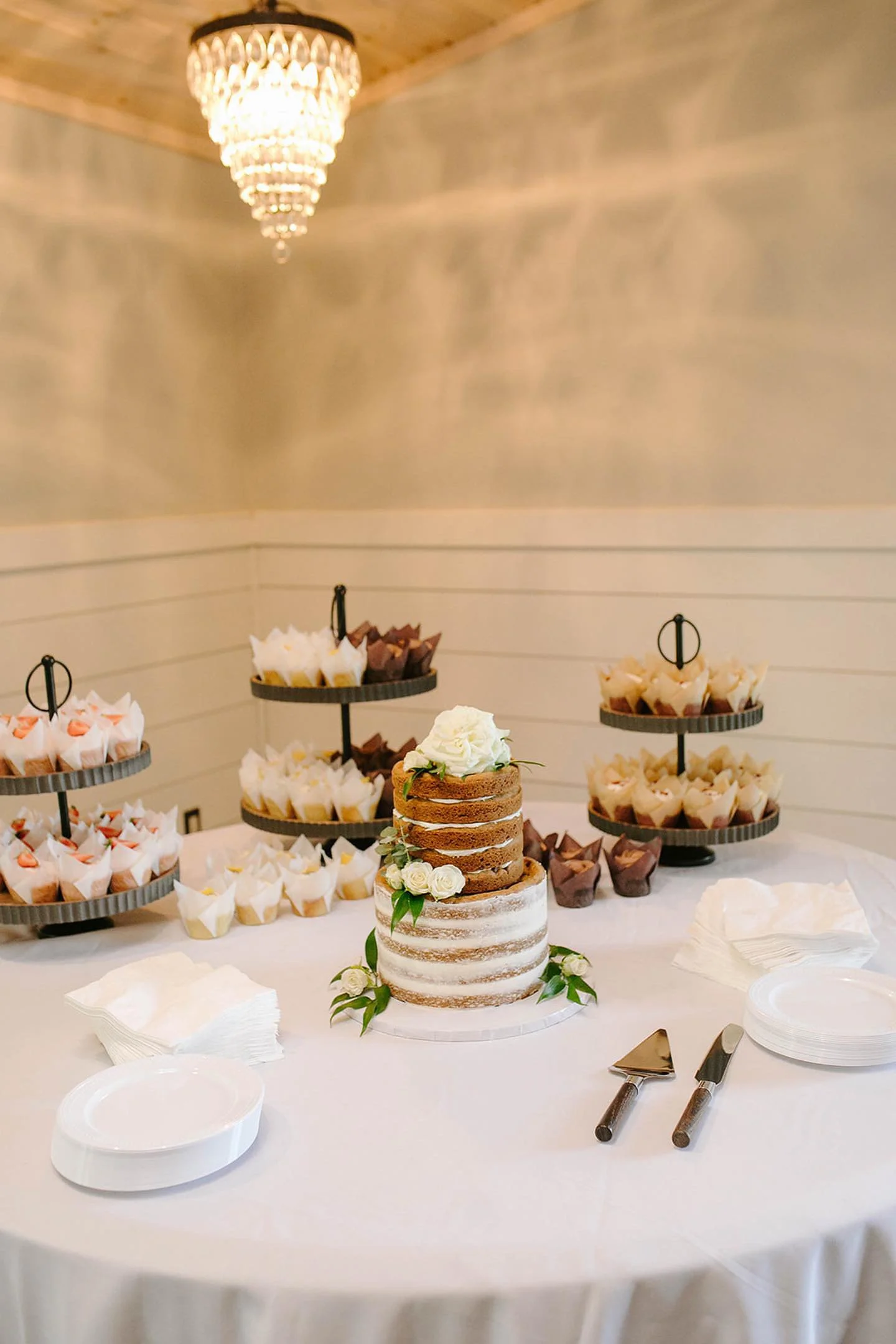 Elegant wedding cake with white and brown frosting decorated with white roses and greenery, placed on a white table with plates, napkins, and cake serving utensils, with cupcake stands holding assorted cupcakes in the background under a chandelier.