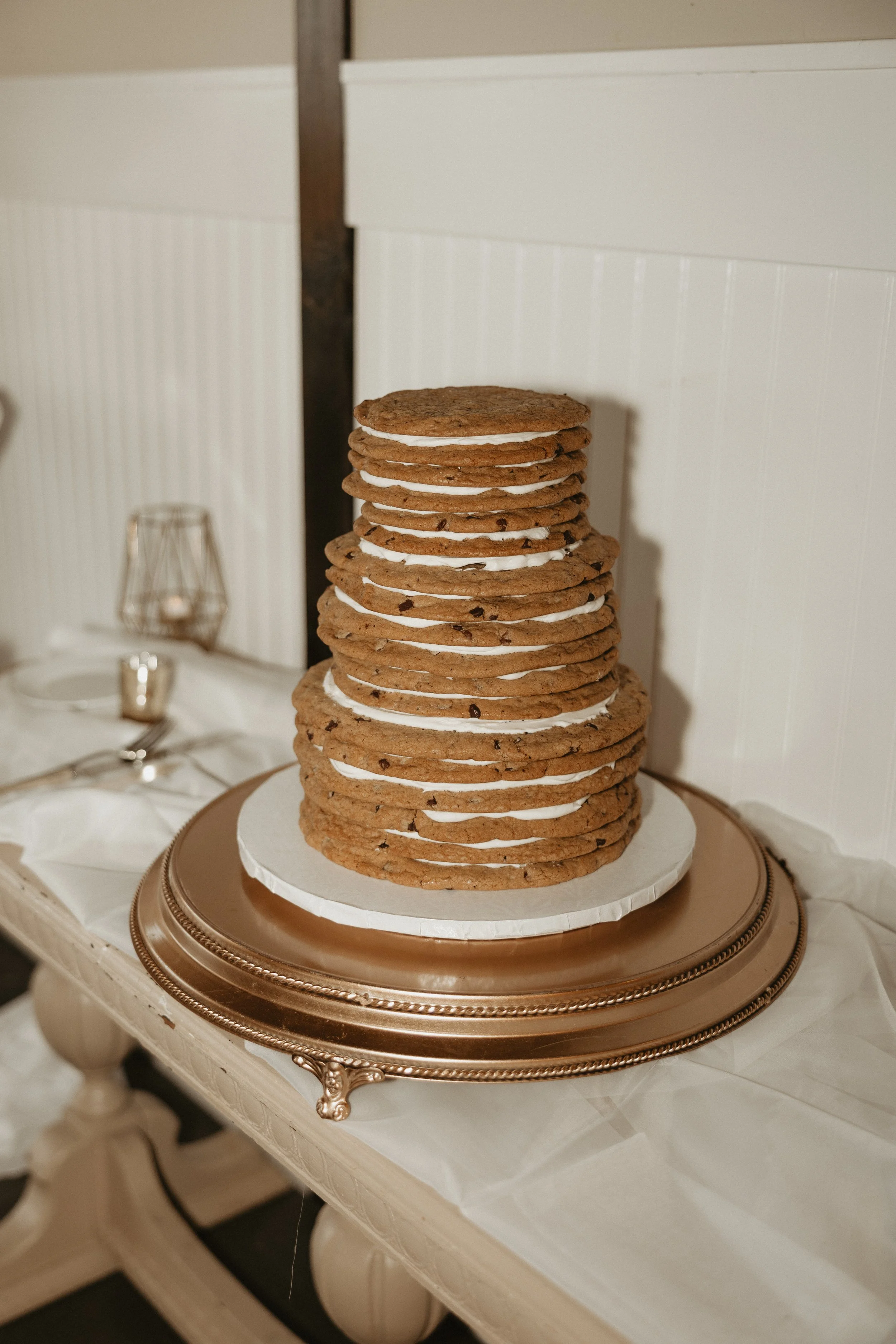 A tall stack of chocolate chip cookies with icing in between each layer, placed on a decorative silver platter on a white table.