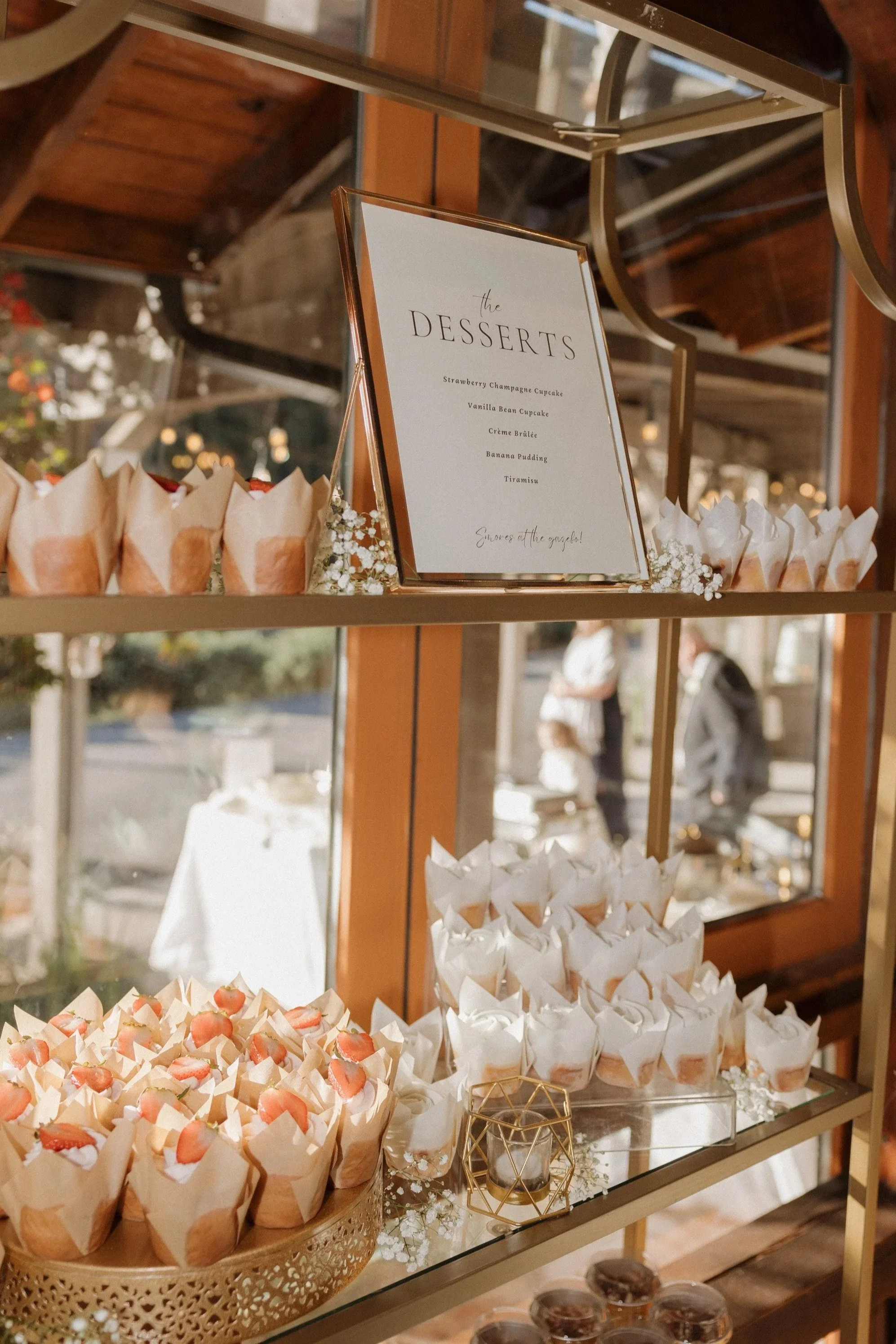 Display of various desserts, including strawberry champagne cupcakes, vanilla bean cupcakes, crème brûlée, banana pudding, and tiramisu, arranged on a gold tiered stand at an event.