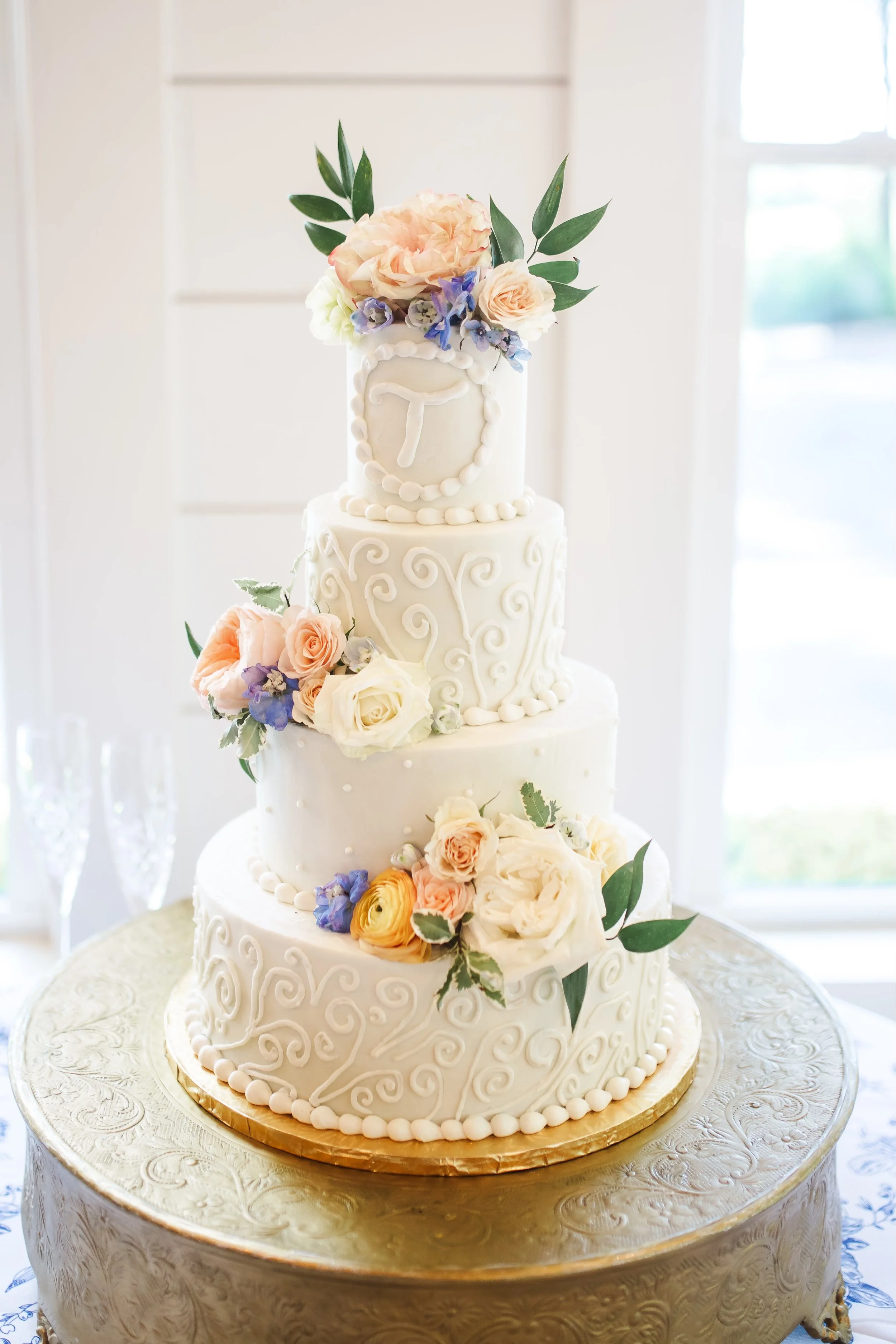 A four-tier white wedding cake decorated with flowers and piping designs, topped with a clock motif and arranged with peach, white, purple, and green foliage, on a gold cake board.
