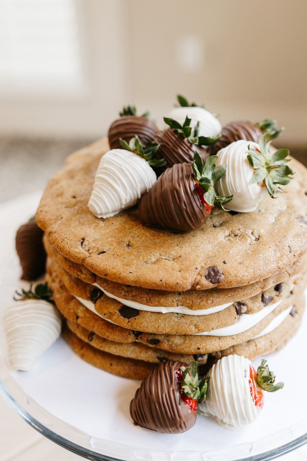 A stack of four chocolate chip cookies decorated with strawberries dipped in white and milk chocolate, with additional strawberries and cookies surrounding the base.