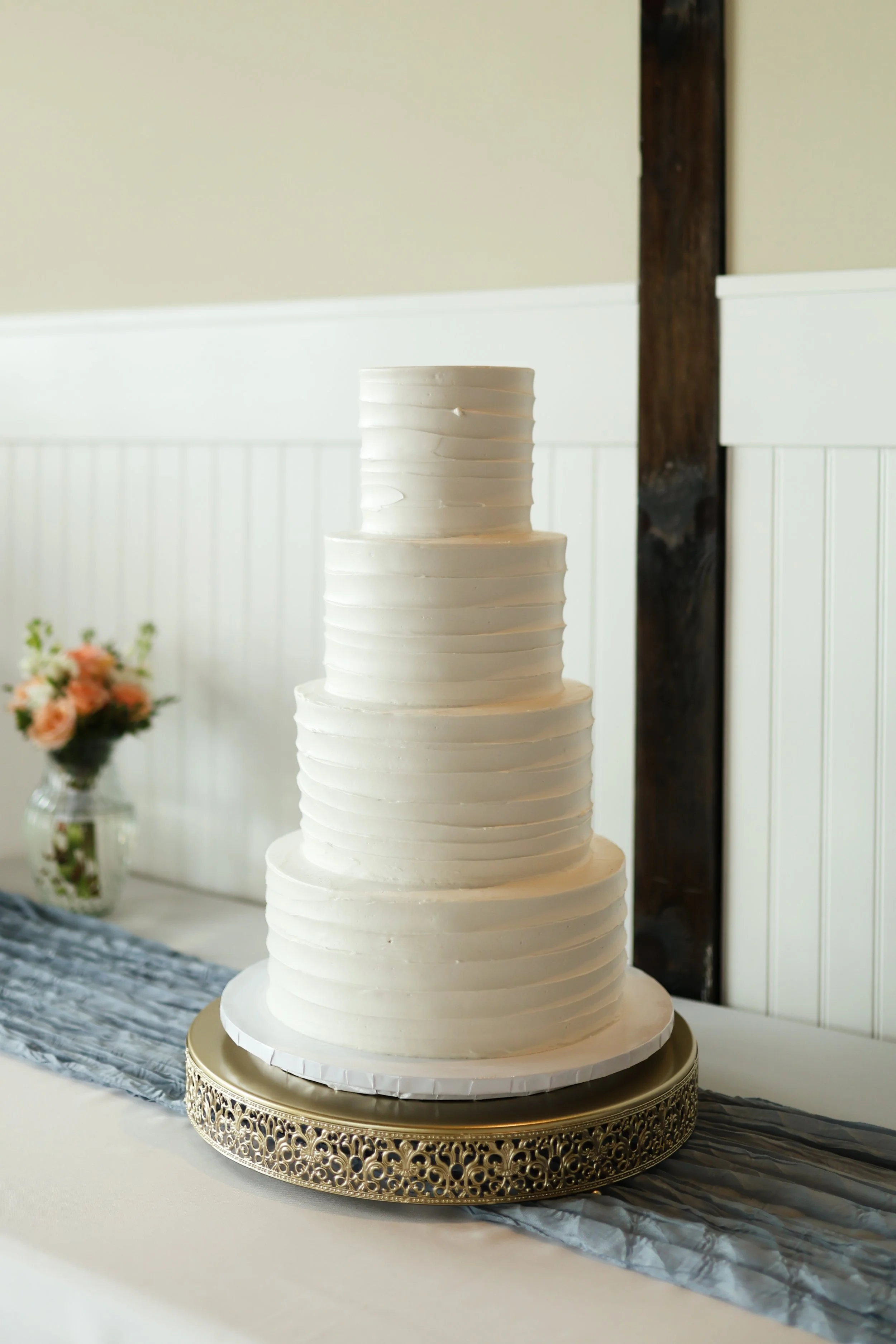 A tall white tiered cake with a modern, textured design, displayed on a decorative gold stand, with a blurred background of a white-walled room and flowers.