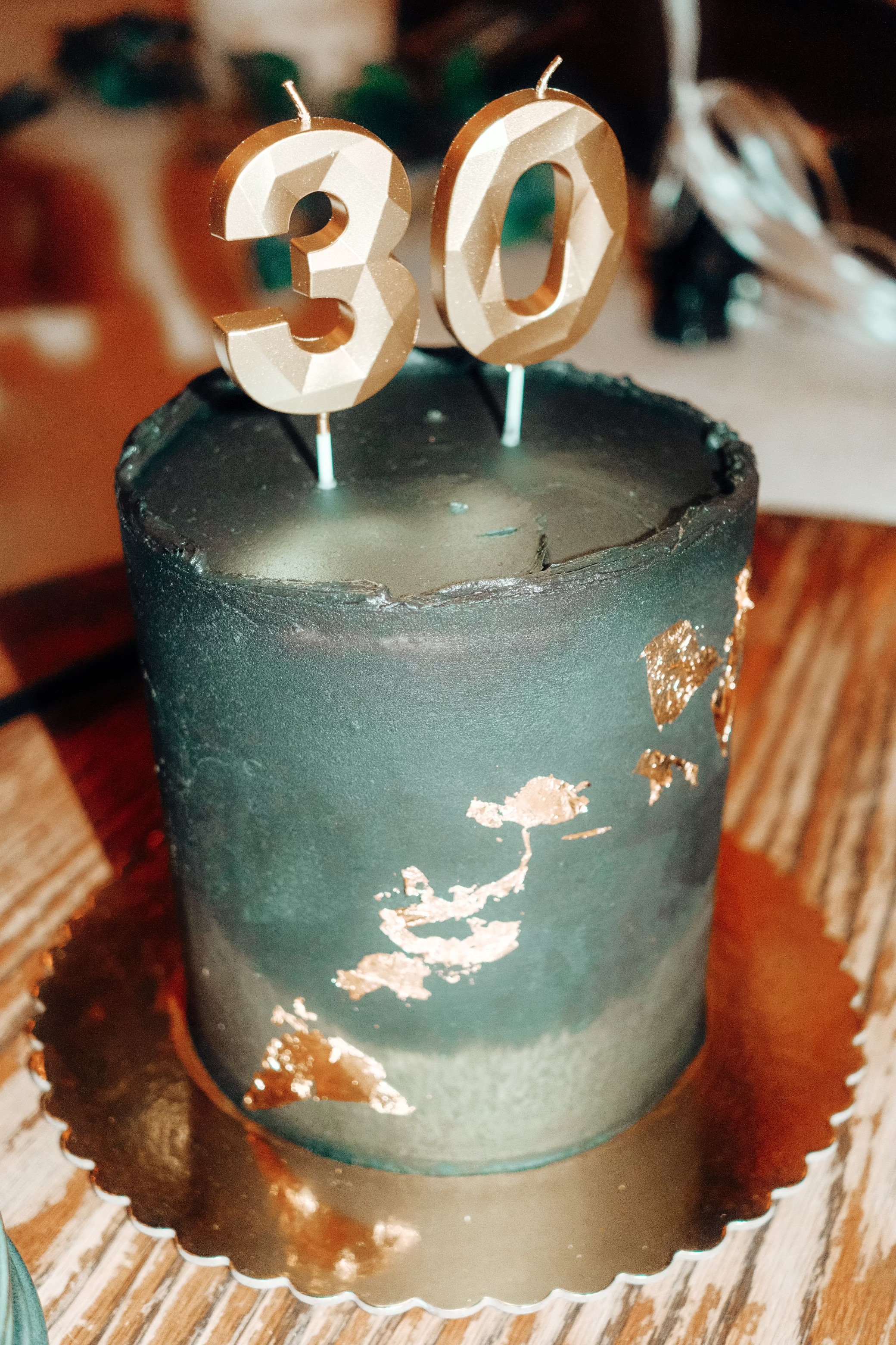 A black birthday cake with gold foil accents on a gold scalloped cake stand, topped with gold number candles displaying '30'.