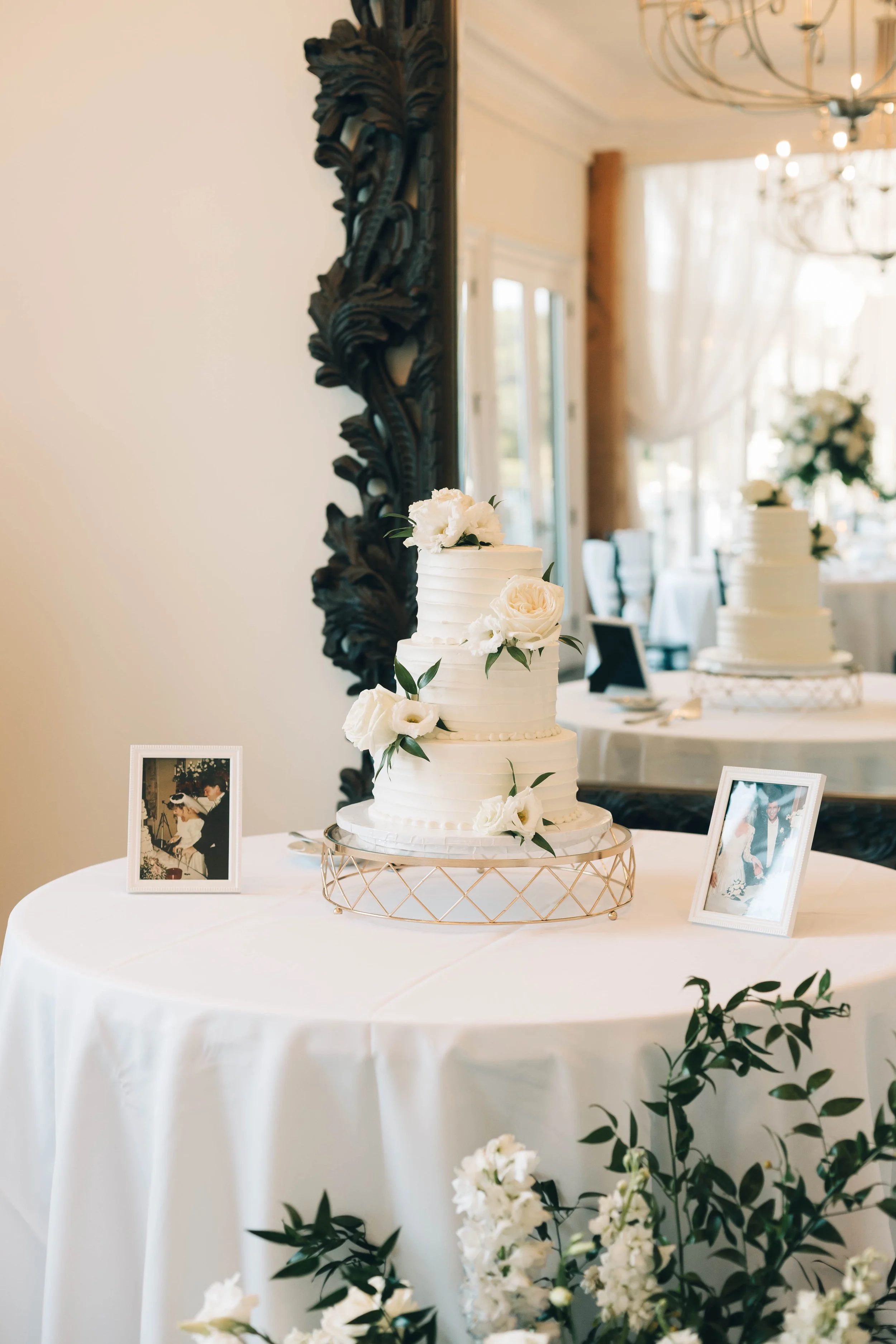A wedding cake with white icing, decorated with white roses and greenery, placed on a round table with white tablecloth, in a decorated event space with flowers, photographs, and elegant lighting.