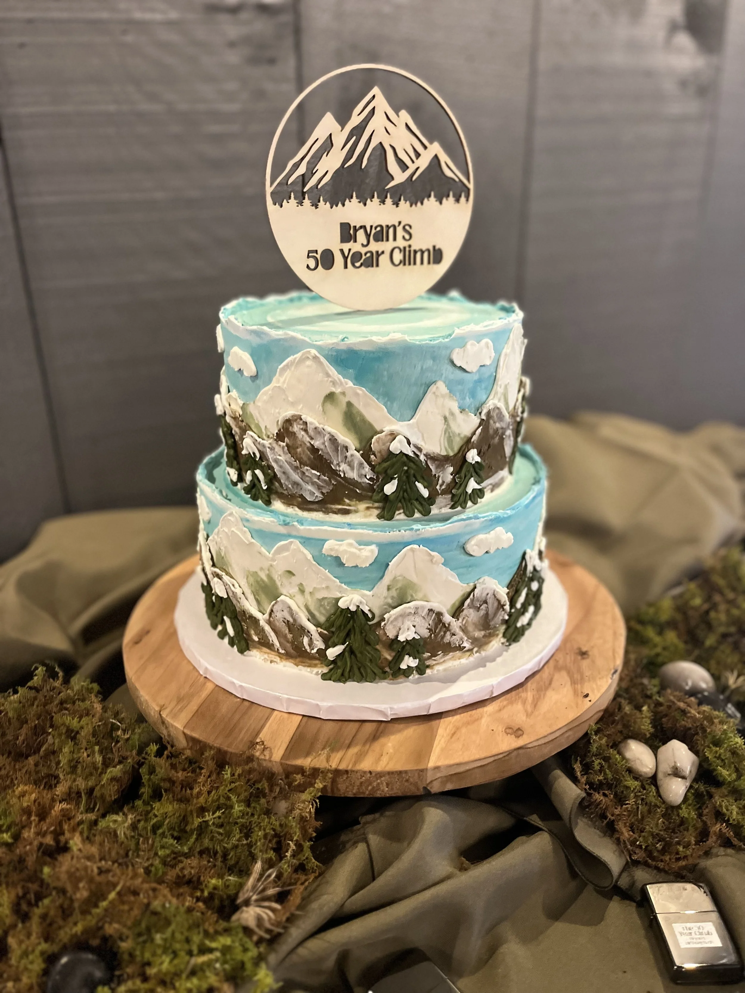 A two-tiered cake decorated with mountains, trees, and clouds, with a wooden cake topper reading "Bryan's 50 Year Climb," on a wooden cake stand. The scene is set on a mossy surface with stones.