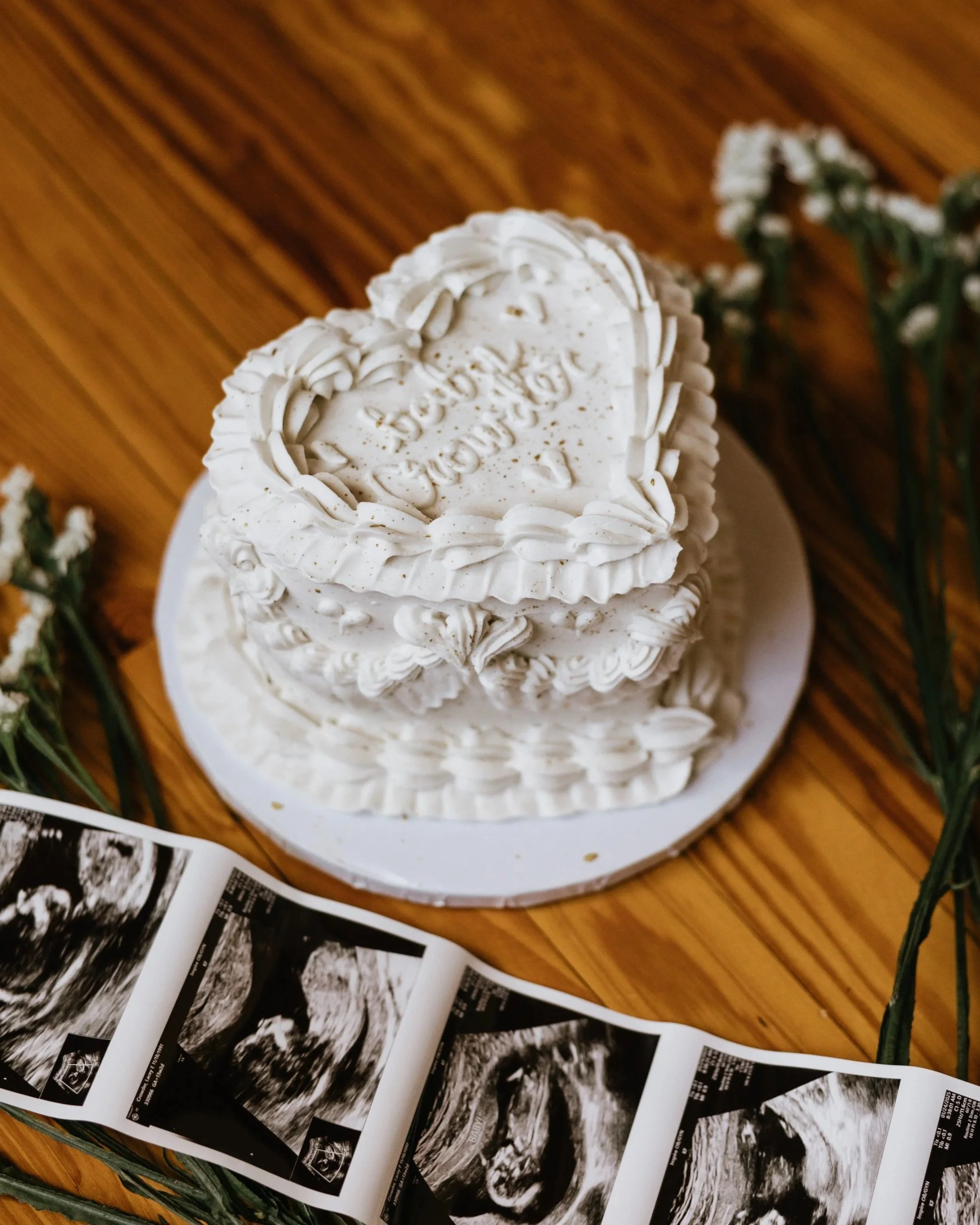 White heart-shaped cake with decorative piping and message, surrounded by printed ultrasound images and white flowers on a wooden surface.