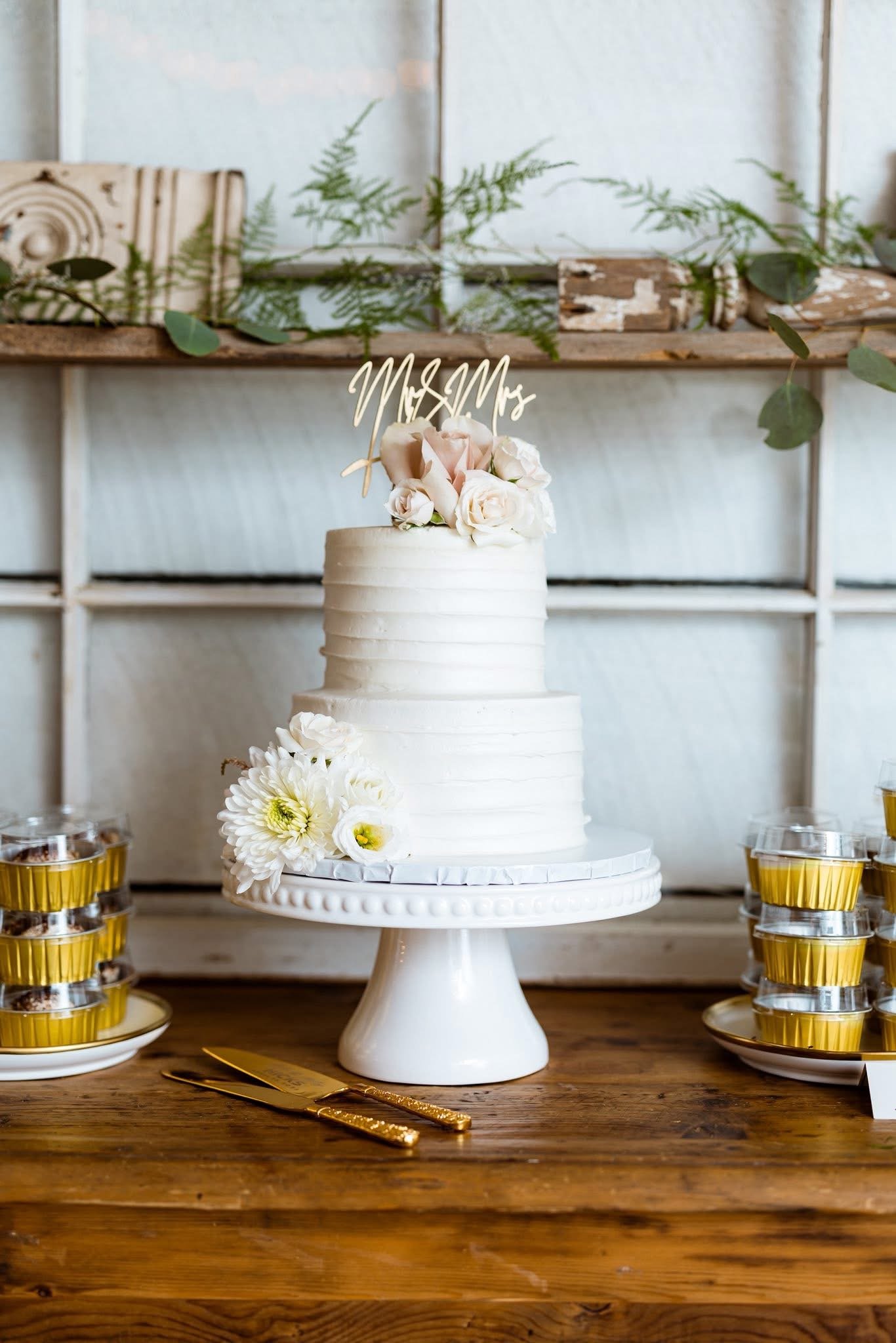 A two-tier white wedding cake with flowers on top and potatoes on the side, placed on a white cake stand on a wooden table, with a "Mr & Mrs" topper.