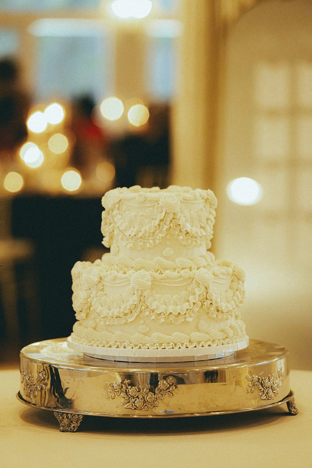 Three-tiered wedding cake with intricate white icing designs, placed on a silver cake stand with floral decorations.