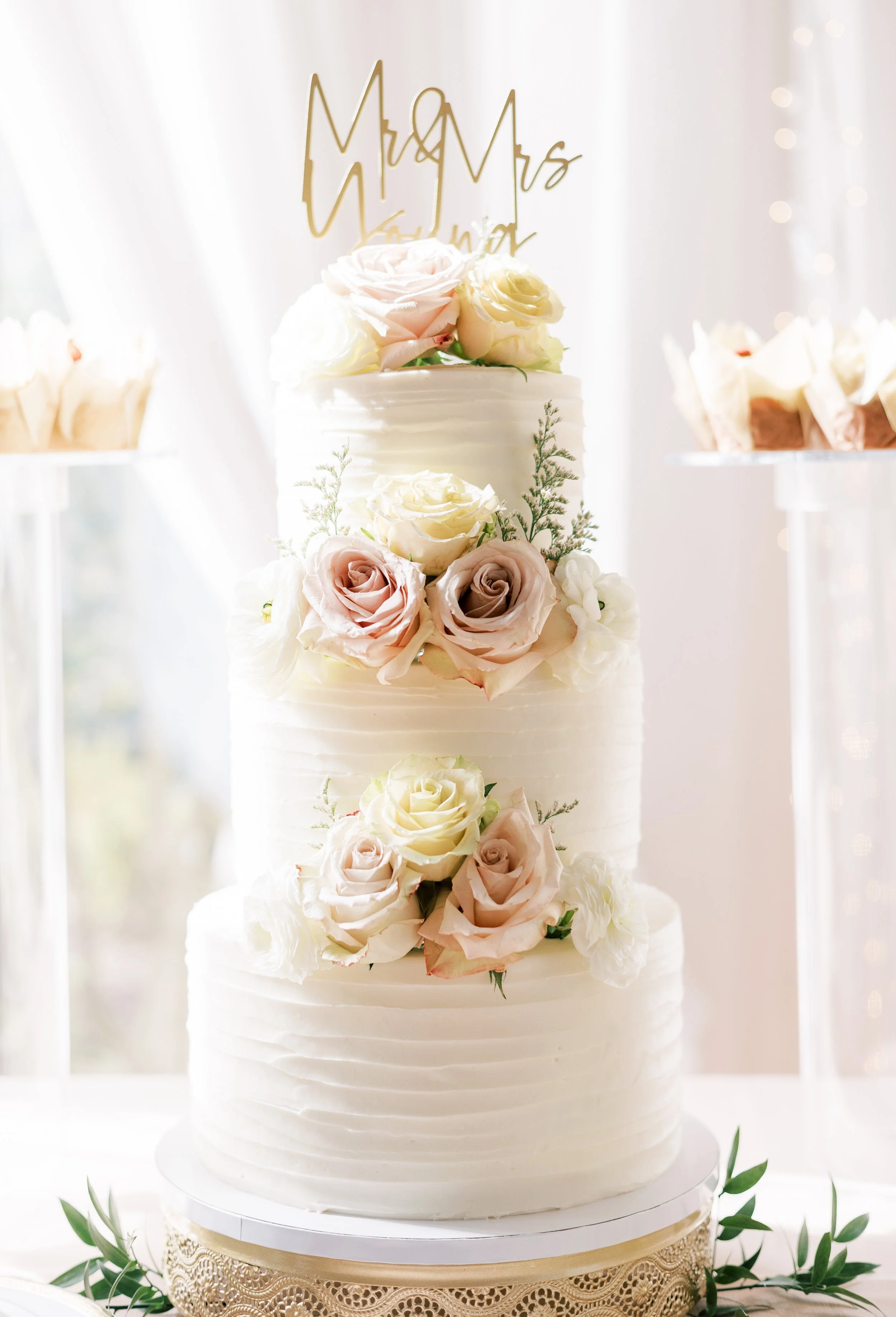A three-tiered white wedding cake decorated with light pink and white roses, with a gold 'Mr & Mrs' cake topper, placed on a decorative gold and white cake stand.